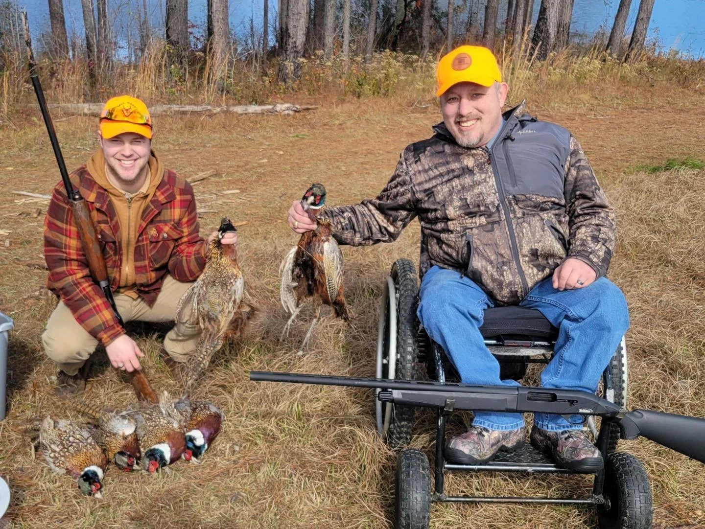 Two men, one in camouflage jacket and wheelchair, the other in plaid jacket, kneeling and sitting on the ground in a field with trees in background, displaying harvested pheasants and ducks, both smiling, one holding a pheasant and the other holding 