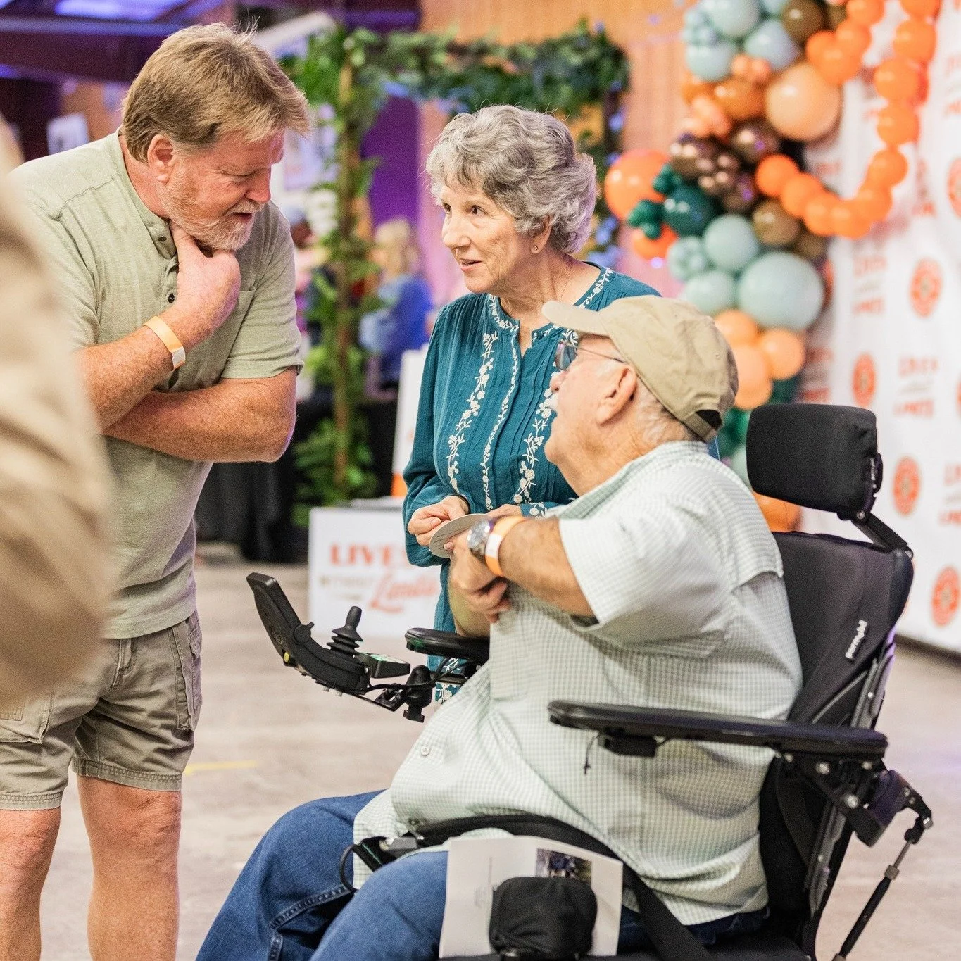 An elderly man in a wheelchair, wearing a baseball cap and a short-sleeved shirt, interacting with a man and a woman at an event. The woman has gray hair and is wearing a teal blouse, while the man is wearing a gray t-shirt. They are in a decorated i