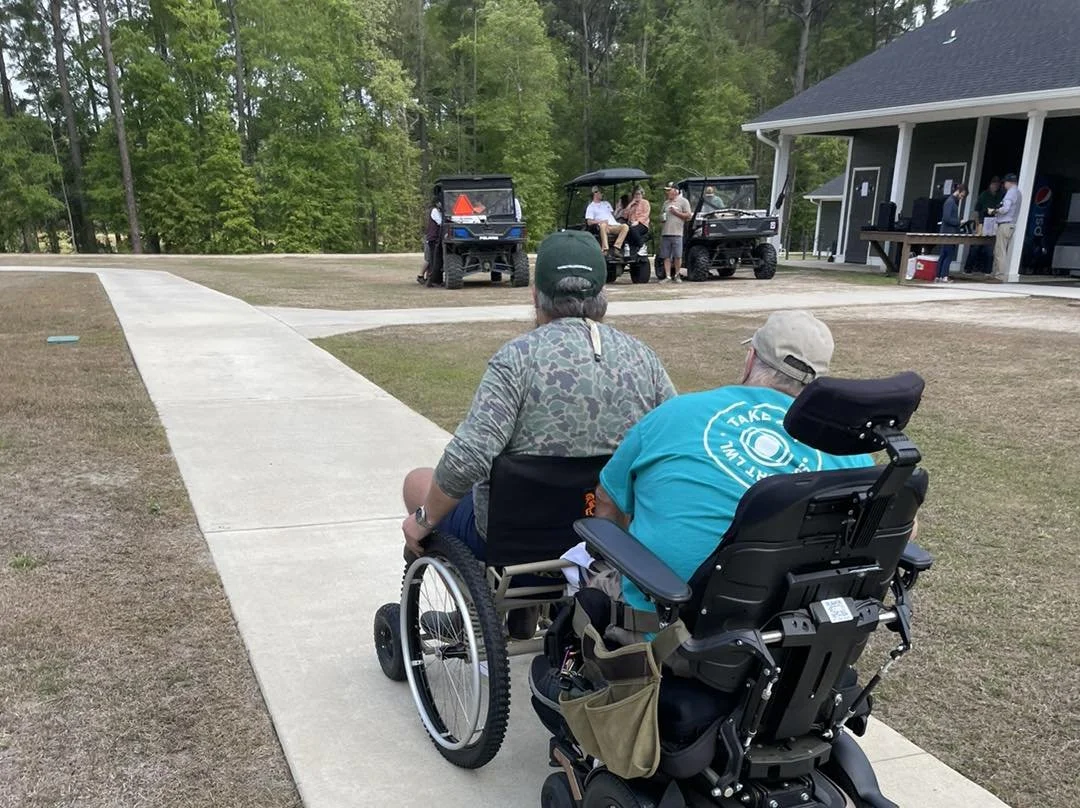 Two people in wheelchairs walking towards a group of people near golf carts and a building with outdoor seating.