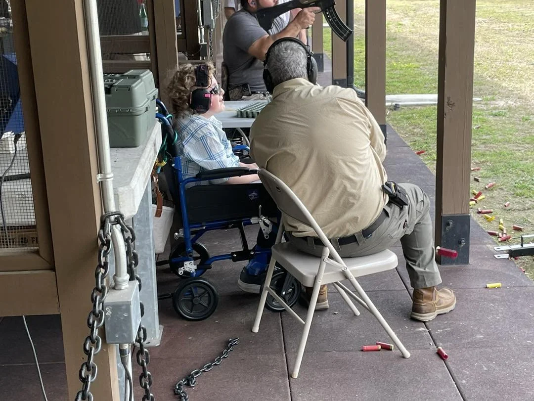 A young girl at a shooting range sitting in a wheelchair, wearing ear and eye protection, aiming a rifle with an instructor nearby. Shotgun shells are scattered on the ground, and others are in use by different shooters.