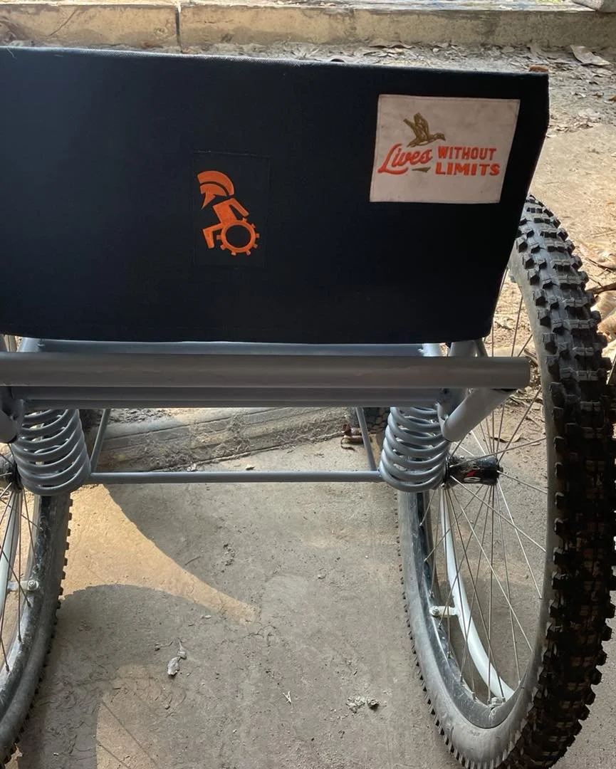 Close-up view of a wheelchair with a black box attached to the back, featuring stickers that say "Lives Without Limits" and a logo in orange. The wheelchair has large, rugged tires and silver metal frame parts.