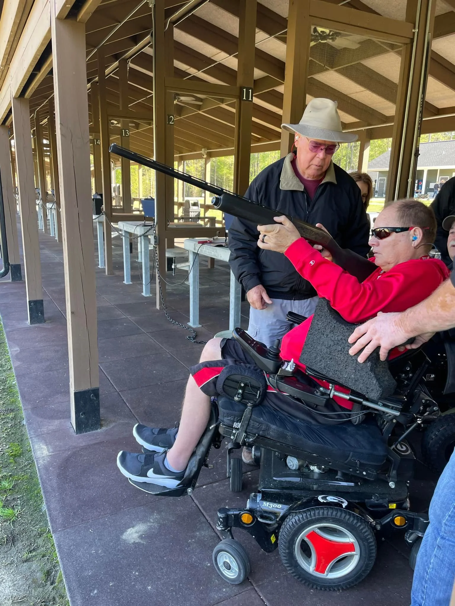 A person in a wheelchair at a shooting range, aiming at a target with assistance from an instructor, under a wooden shelter with numbered lanes.
