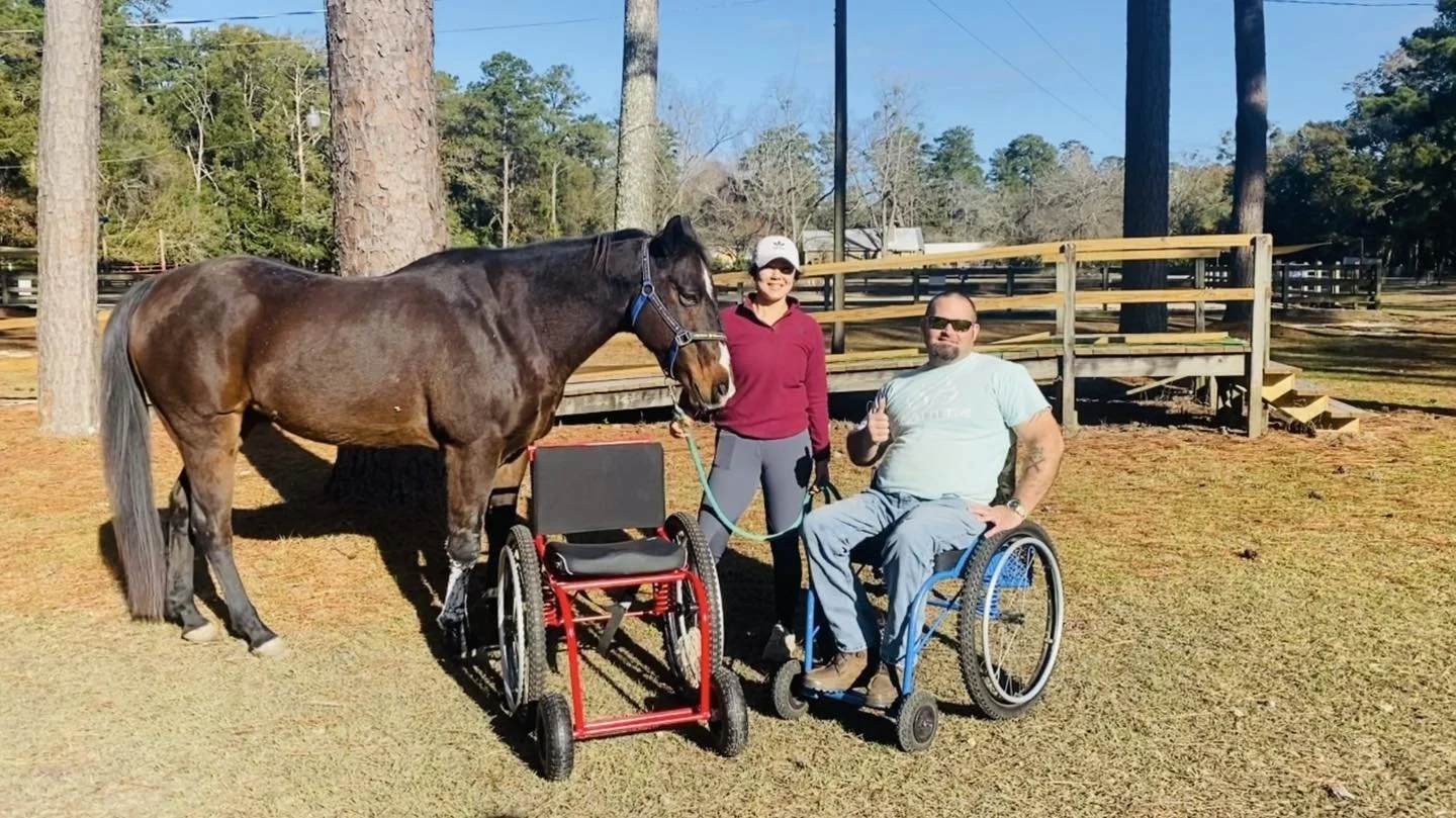 A woman and a man in a wheelchair standing outdoors with a horse and two wheelchairs, in a wooded area under clear blue skies.