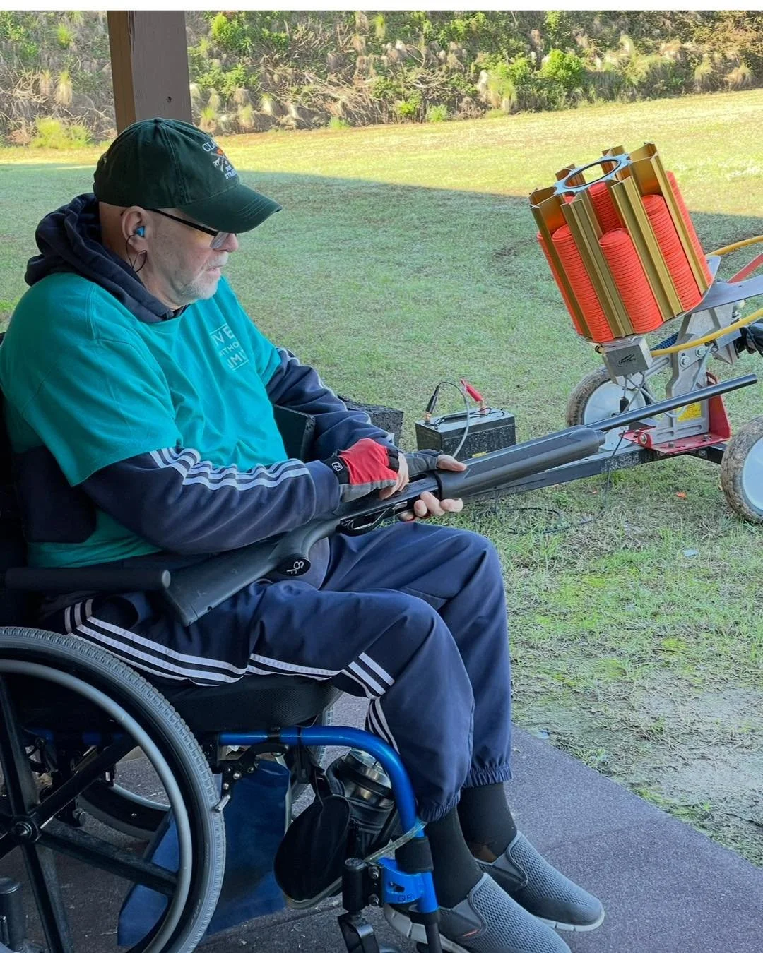 A man in a wheelchair aiming a firearm fitted with a large launcher on a grassy outdoor shooting range.