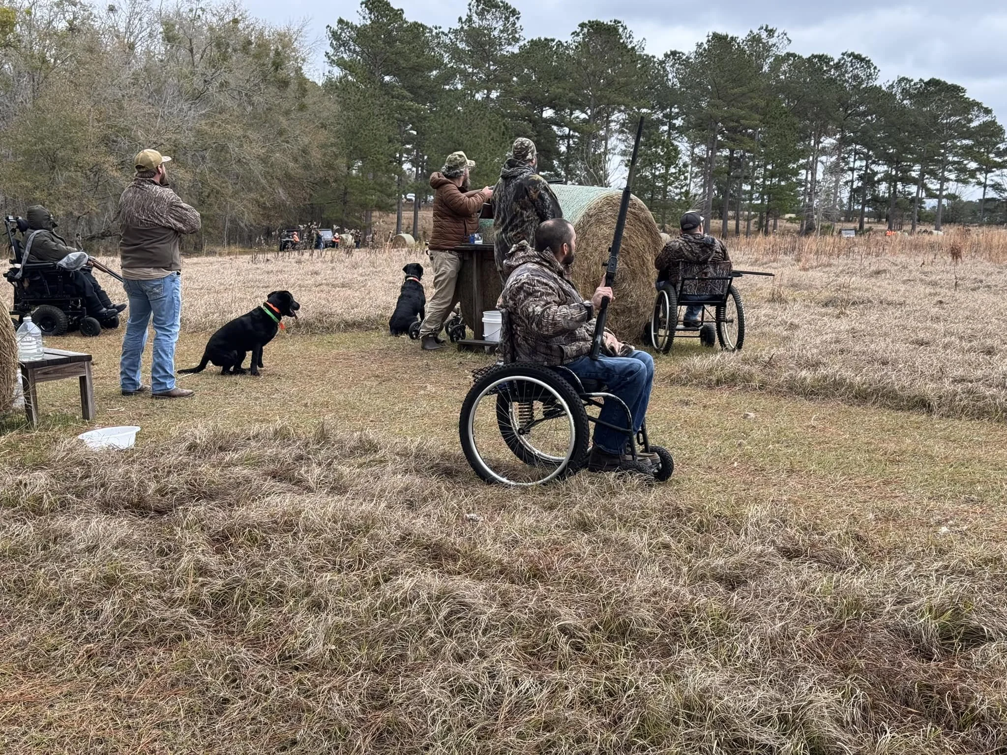 People in camouflage and outdoor gear standing and sitting in a grassy field, with some using wheelchairs, watching or participating in outdoor activity, with trees in the background.