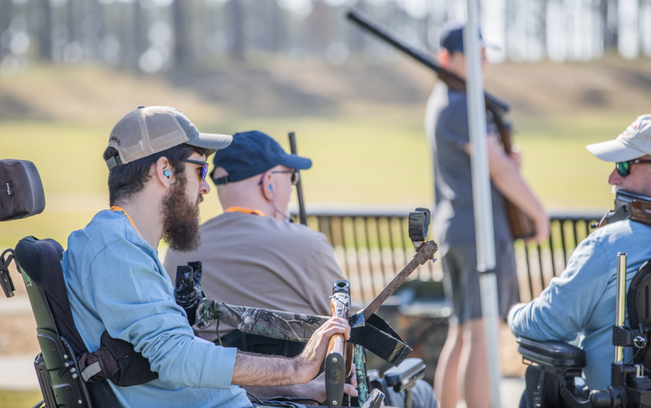 Three men in wheelchairs participating in a shooting activity outdoors, with a person in the background holding a rifle.