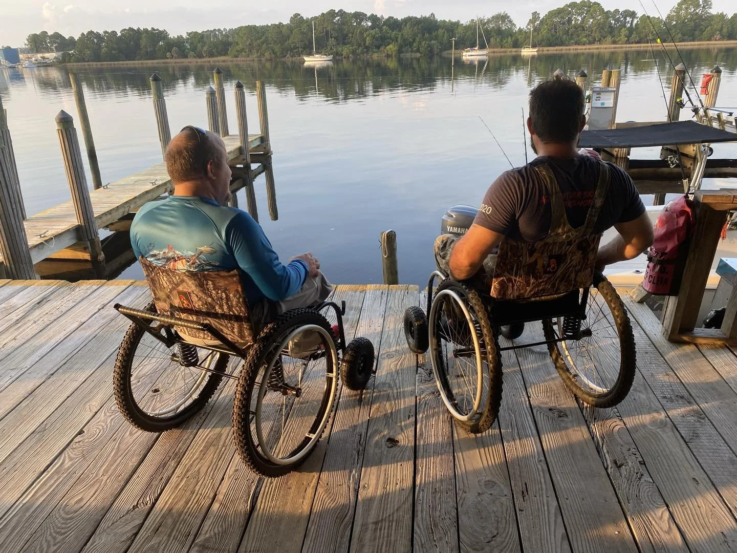 Two men in wheelchairs sit on a wooden dock overlooking a calm body of water with boats and trees in the distance.