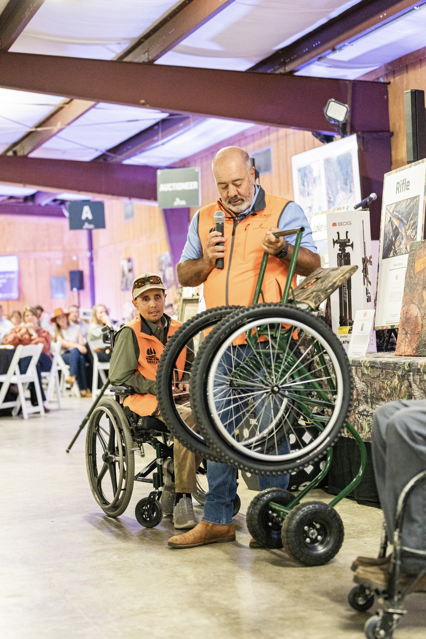 Two men, one in a wheelchair and the other standing, at an indoor event. The standing man is holding a microphone and looking at a display with outdoor equipment, while the seated man looks at the camera. Both are wearing orange vests. The background shows an audience seated in chairs and signs indicating different auction areas.