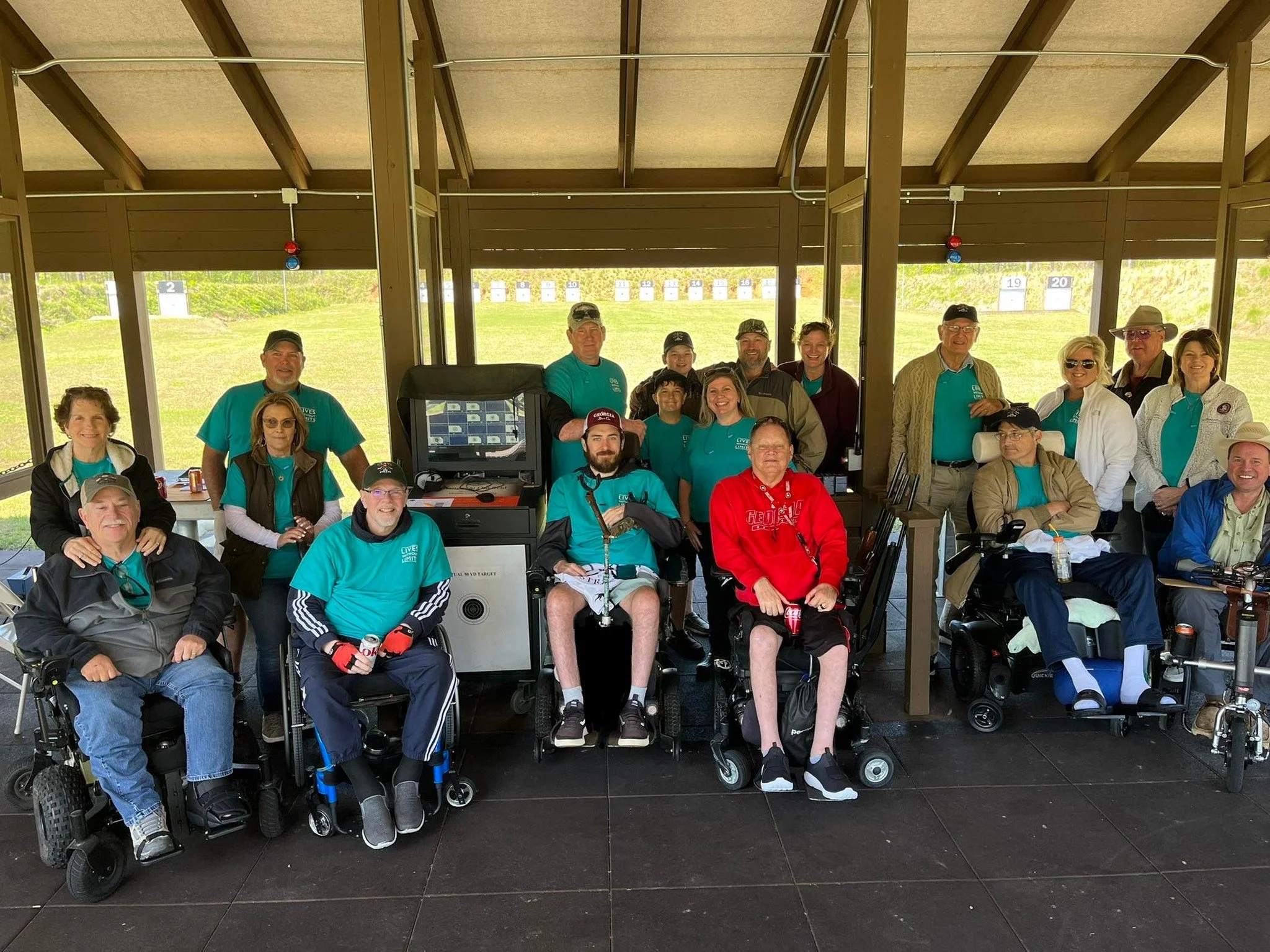 Group of people at a shooting range, some in wheelchairs, posing for a photo indoors with targets visible outside in the background.