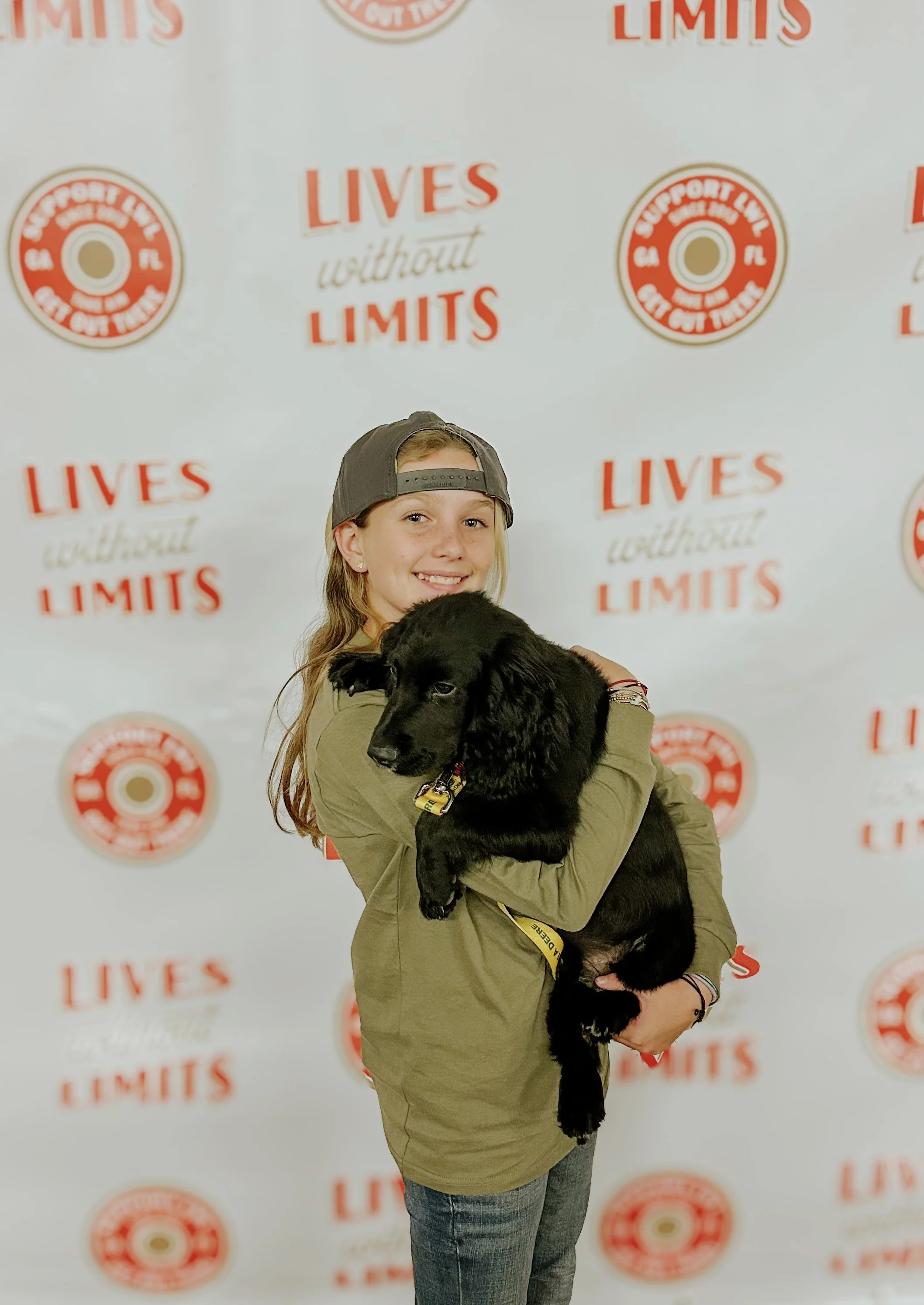Smiling young woman in a backward gray cap, khaki shirt, and jeans holding a black puppy with floppy ears against a backdrop with red and white text and logos reading 'LIVES without LIMITS' and 'Support Life and Gift Limit'.