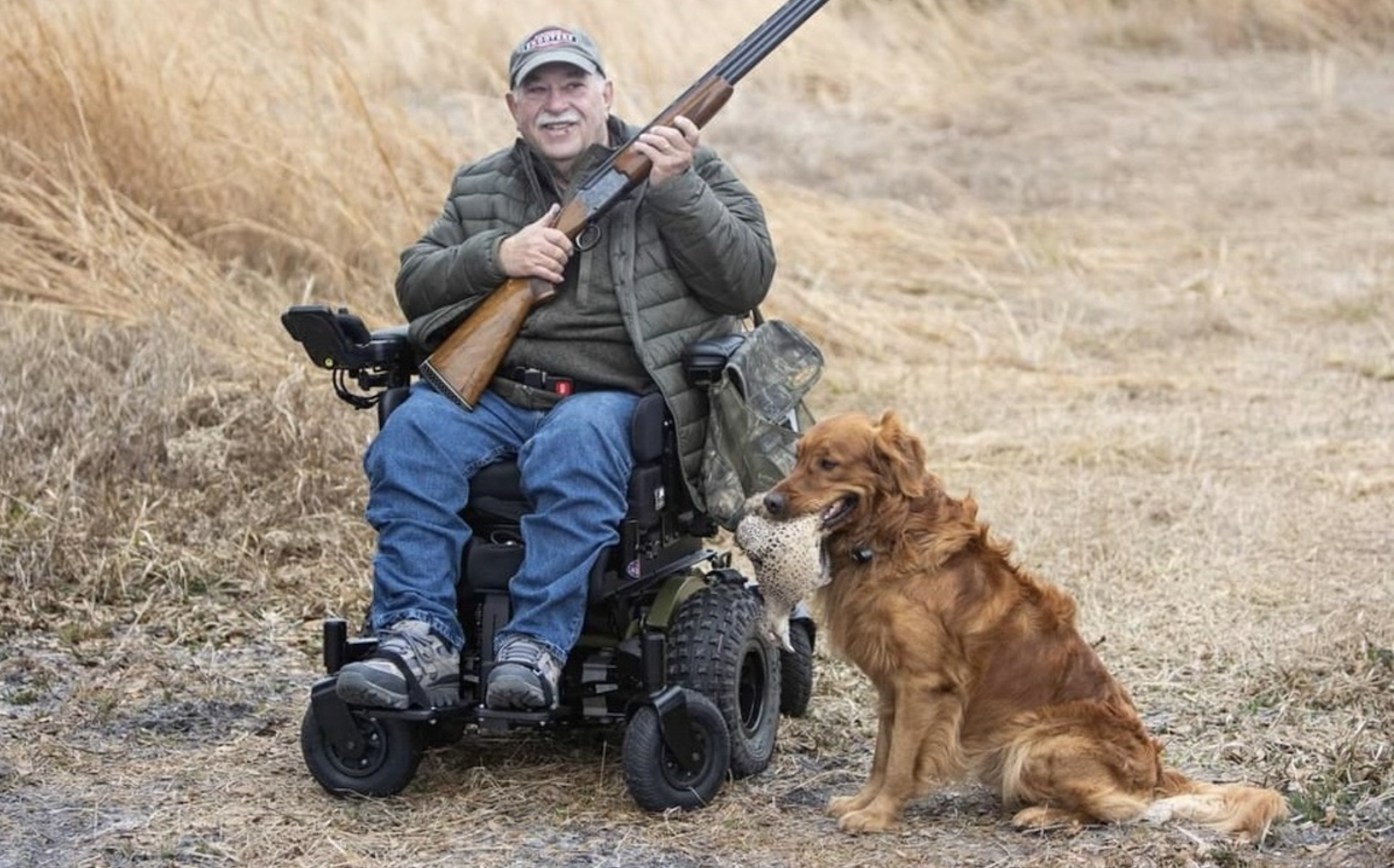 Man in a motorized wheelchair outdoors with a golden retriever dog holding a bird in its mouth.