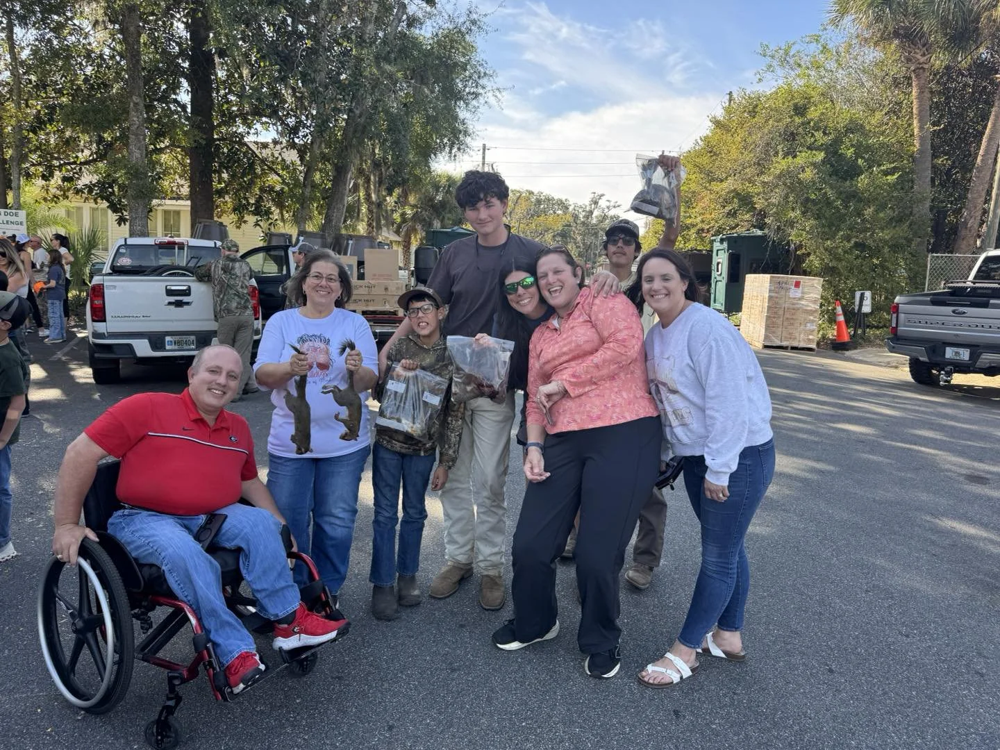 Group of smiling people, including children and adults, gathering outdoors in a parking lot, some holding boxes and bags of what appear to be small animals or bait, with trucks and trees in the background.