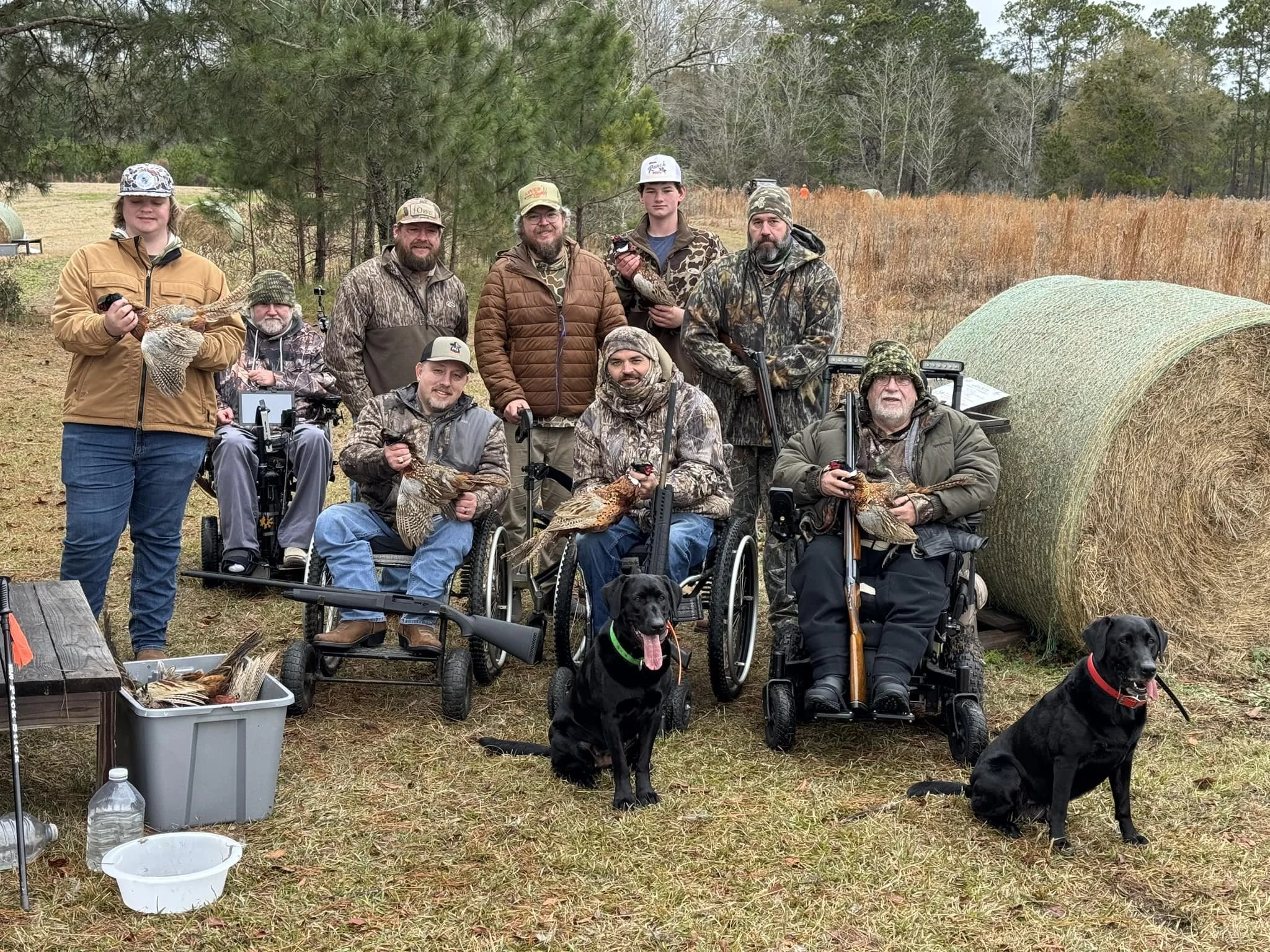 Group of hunting enthusiasts, some with disabilities, posing outdoors with harvested game and hunting gear, including rifles and dogs, near a large hay bale.