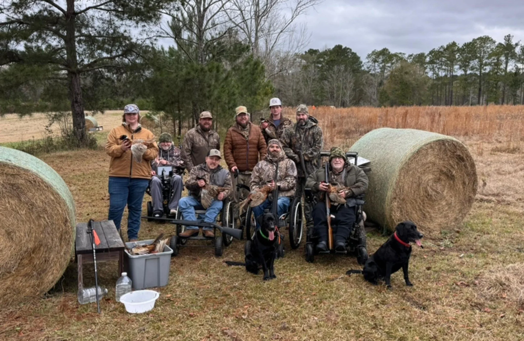 A group of people, including some in wheelchairs, are gathered outdoors in a field with hay bales. They are dressed in camouflage or outdoor clothing, and some are holding hunting equipment. Two dogs are sitting in front of the group. The background 