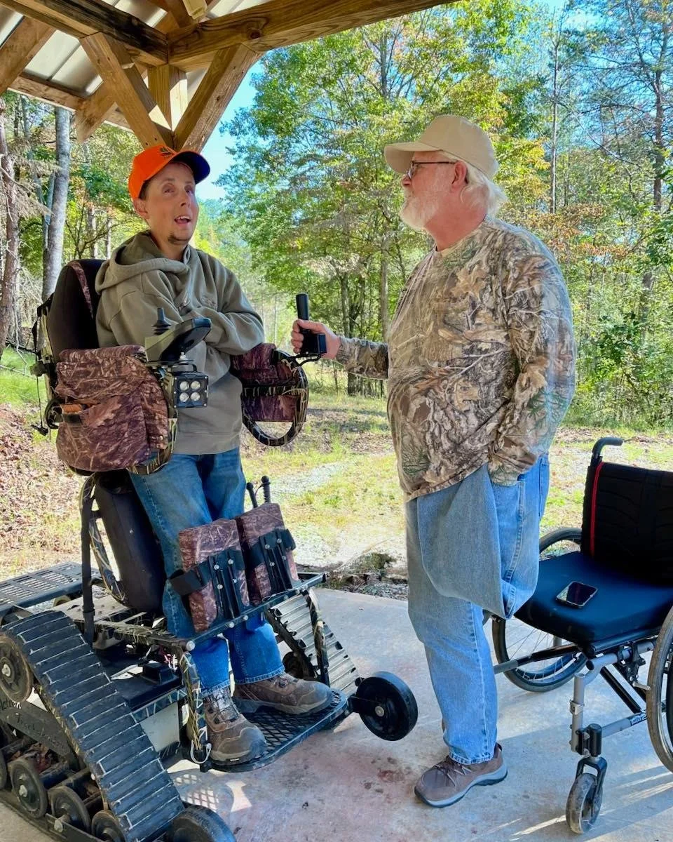 An elderly man with a beard wearing a camouflage long sleeve shirt, jeans, brown shoes, and a beige cap interviewing a woman in a motorized wheelchair. The woman is wearing a gray hoodie, an orange cap, blue jeans, and brown boots. They are outdoors 