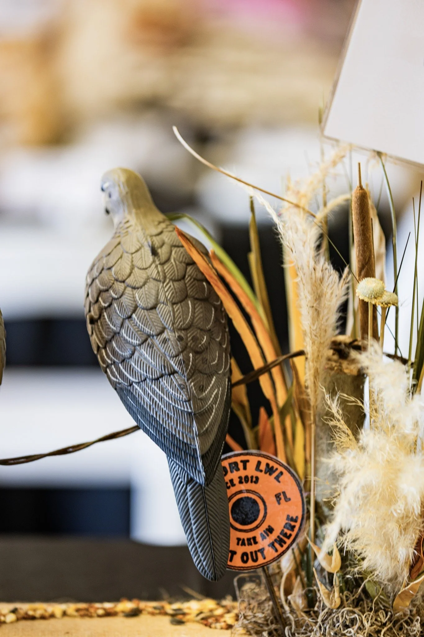 Decorative bird figurine perched on a branch surrounded by dried plants and a small orange sign reading 'SPORT LWL FL 2019 FL TABLE AND TAKE AND PUT OUT THERE'.