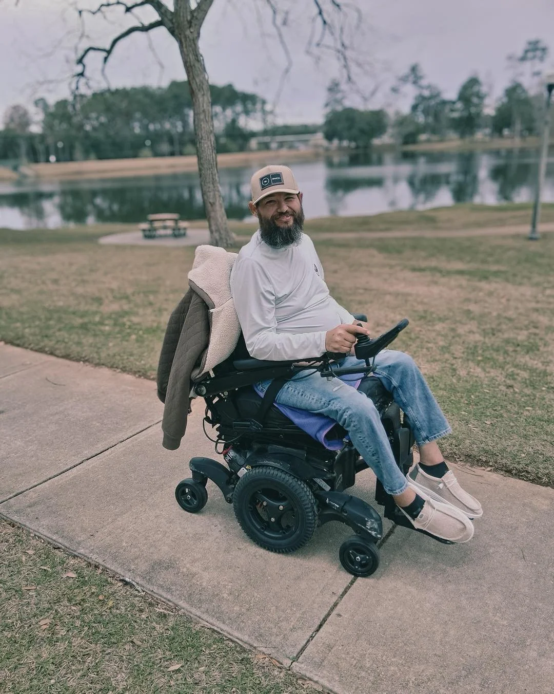 Man with a beard sitting in a motorized wheelchair near a lake in a park, holding a remote control device, with a jacket draped over the wheelchair and a landscape of trees and water in the background.