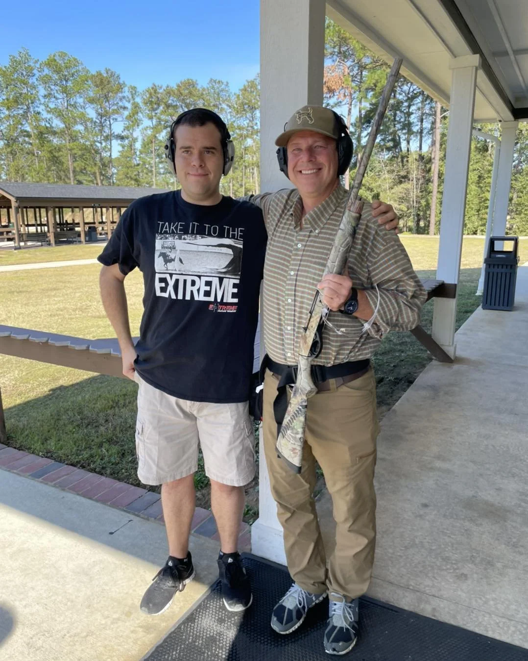 Two men standing outdoors on a porch, one holding a rifle, both wearing ear protection and smiling.