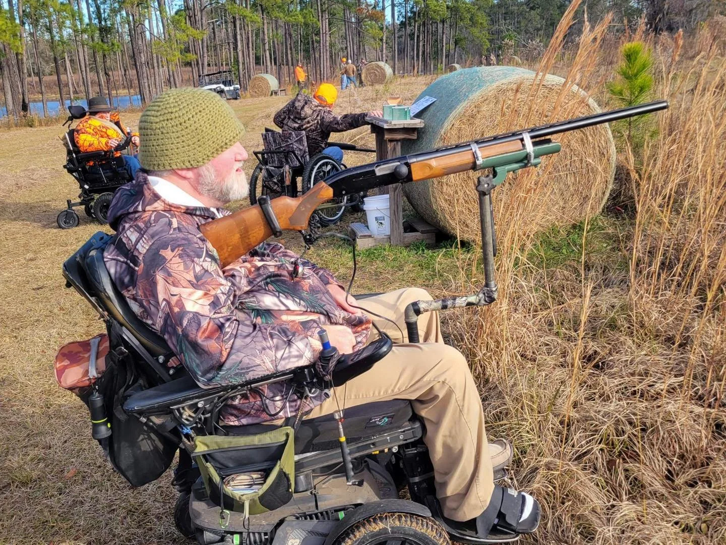 A man in a wheelchair aiming a shotgun while shooting clay targets in an outdoor field with tall grass and hay bales, other people and trees in the background.