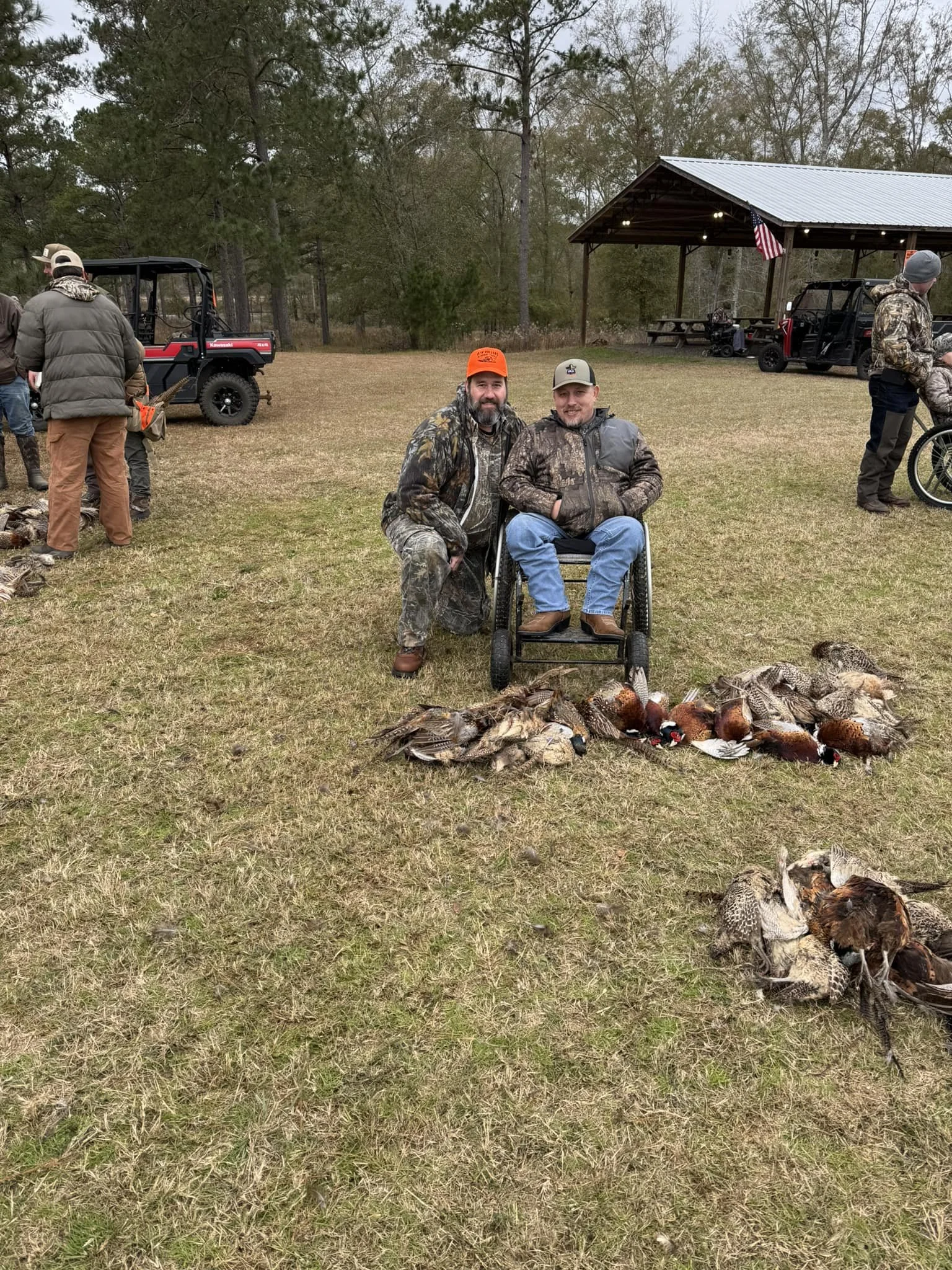 Two men, one in a wheelchair, pose in front of a display of various hunted ducks, with a group of people and utility vehicles in the background at an outdoor hunting area.