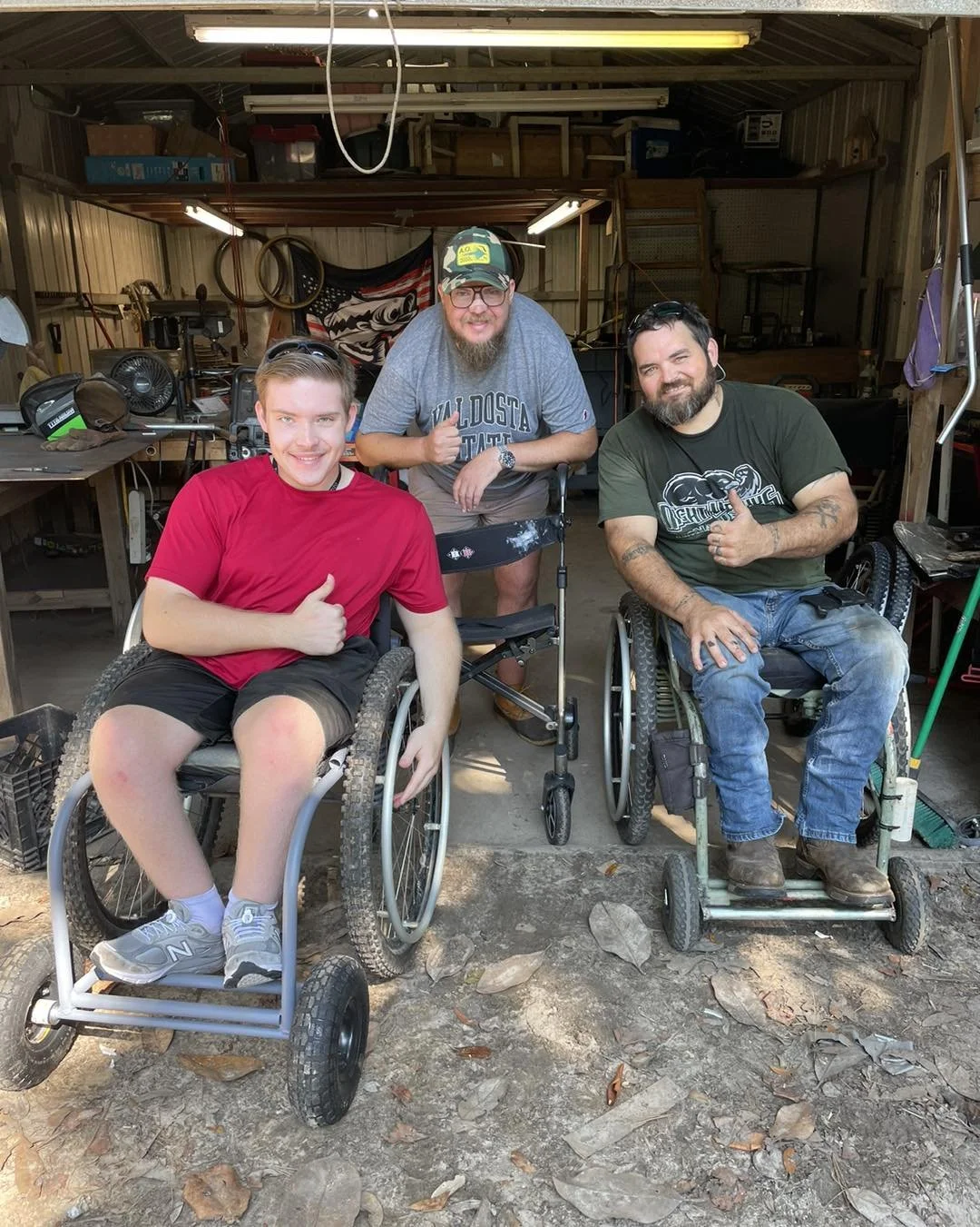 Three men, two in wheelchairs and one standing behind, smiling and giving thumbs-up in a garage workshop.