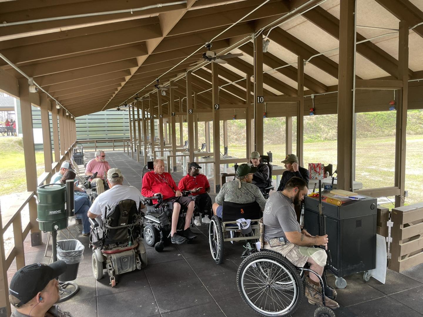 A group of people, some in wheelchairs, sitting under a covered outdoor area, possibly at a shooting range or similar facility.