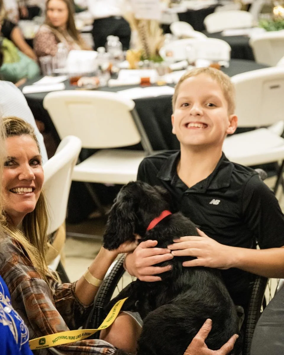A smiling boy holding a black puppy with a red collar at a social gathering or event.