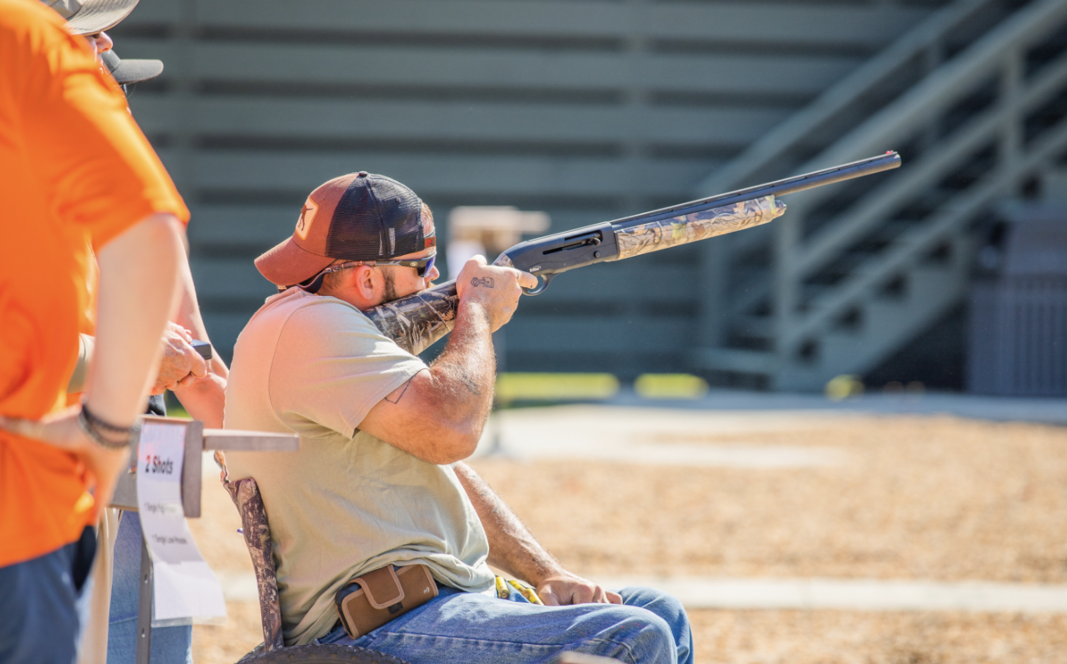 A man seated in a wheelchair aiming a shotgun during a shooting competition, with a person in an orange shirt nearby, outdoors at a shooting range.