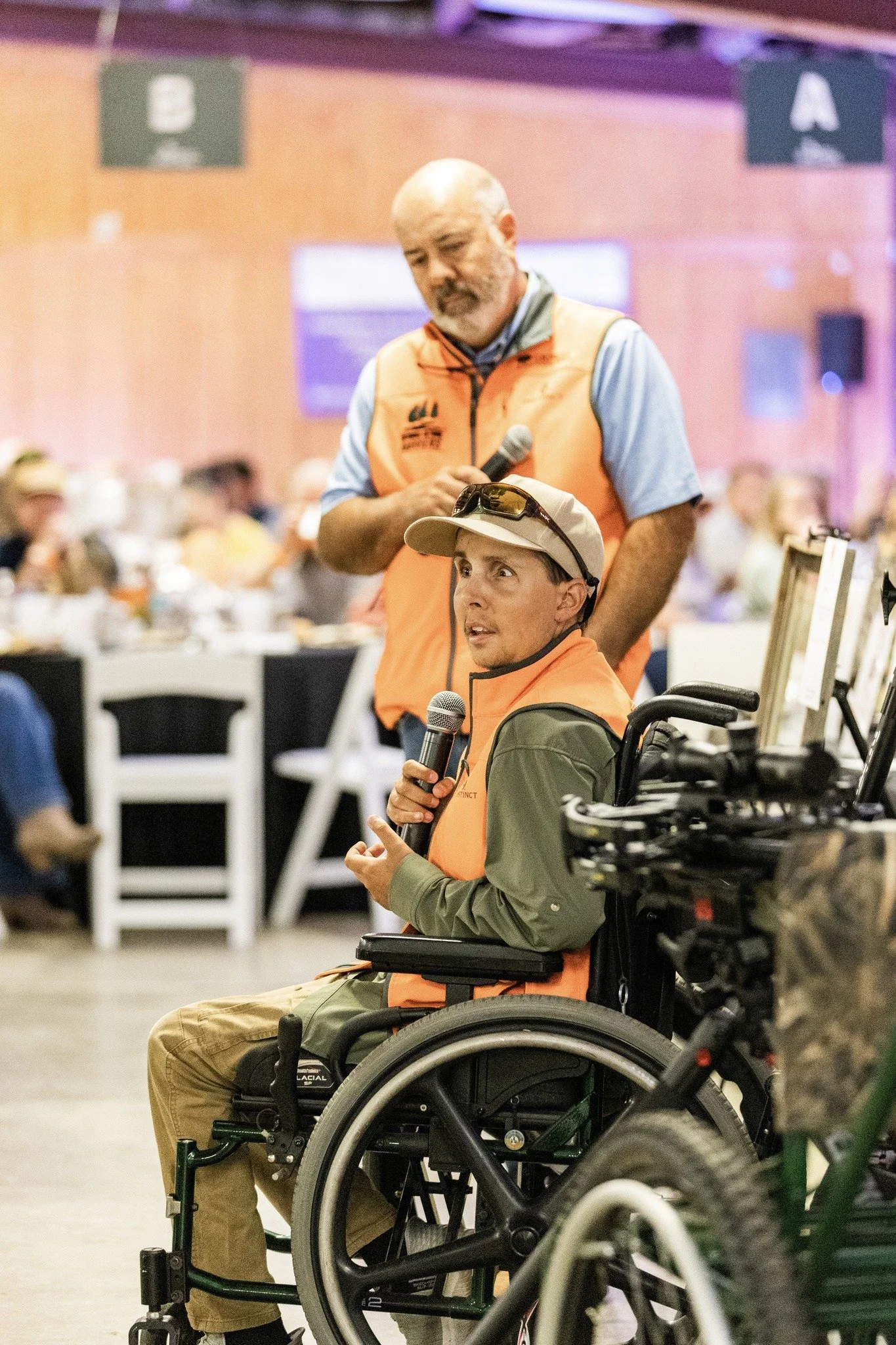 Two men in orange vests during an indoor event; one is standing with a microphone and the other is sitting in a wheelchair, also holding a microphone, with people seated at tables in the background.
