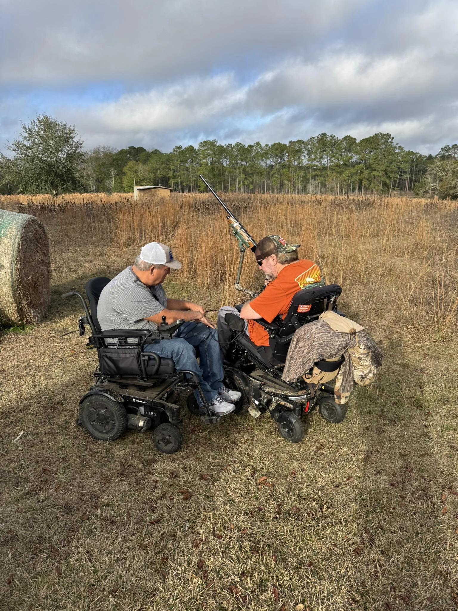Two men in power wheelchairs outdoors in a field with tall grass, sitting closely together, with one man adjusting or working on something as they converse, under a partly cloudy sky.
