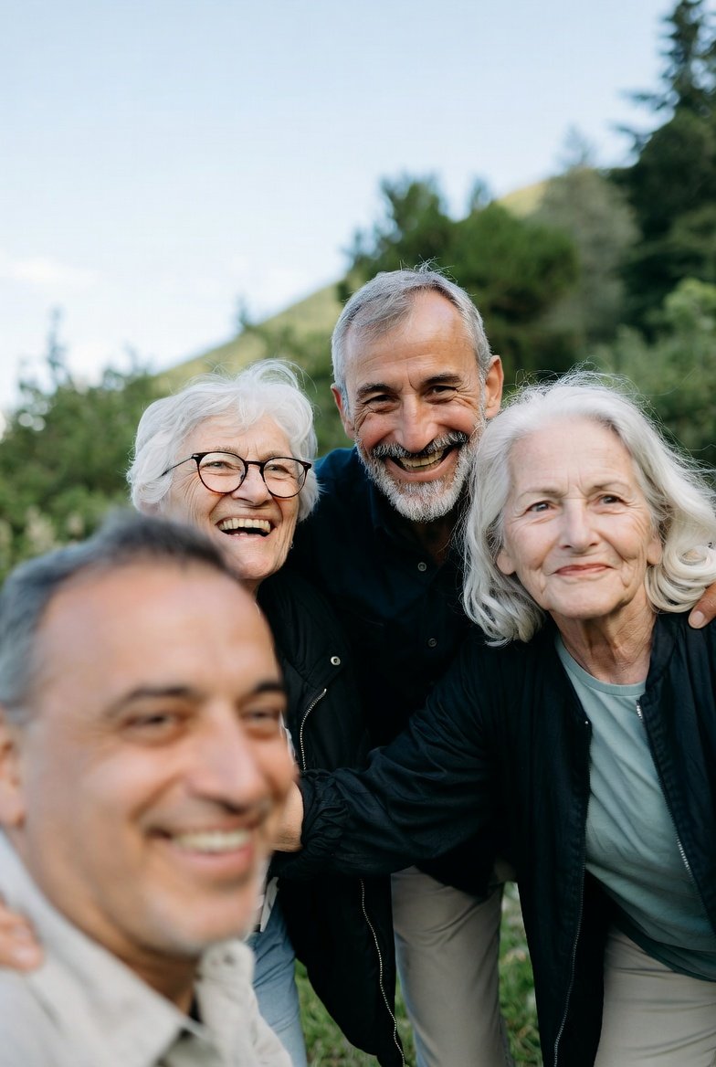 Smiling elderly adults and middle-aged man taking a group selfie outdoors near green hills and trees.