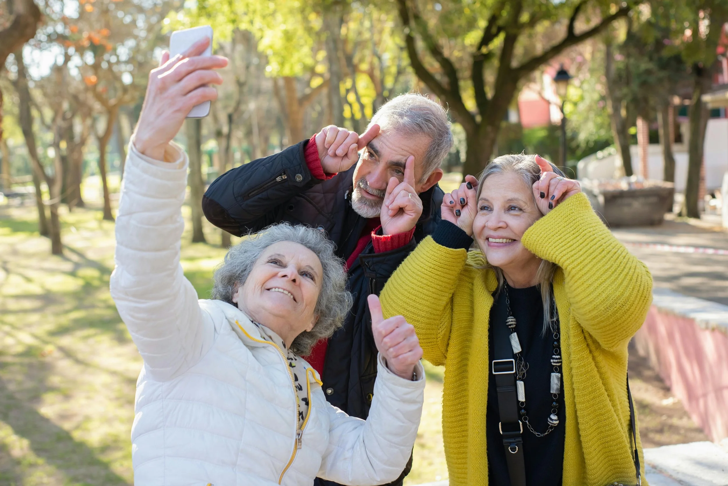 Four elderly friends taking a selfie together in a park on a sunny day, smiling and making playful gestures.