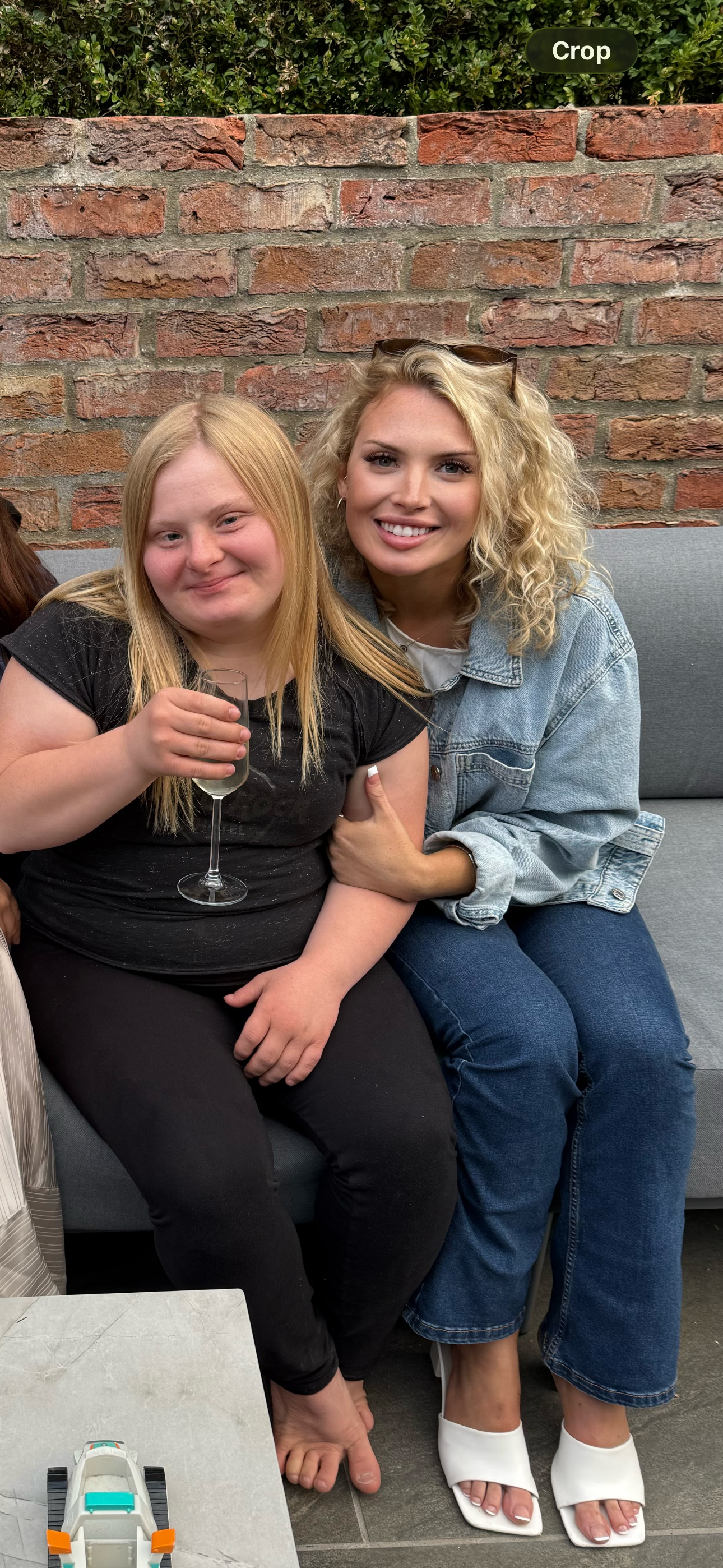Two women sitting on a gray couch against a brick wall, smiling at the camera. One woman, with blonde curly hair, is wearing sunglasses on her head and a light blue denim jacket. The other woman, with straight blonde hair, is holding a glass of what appears to be water.