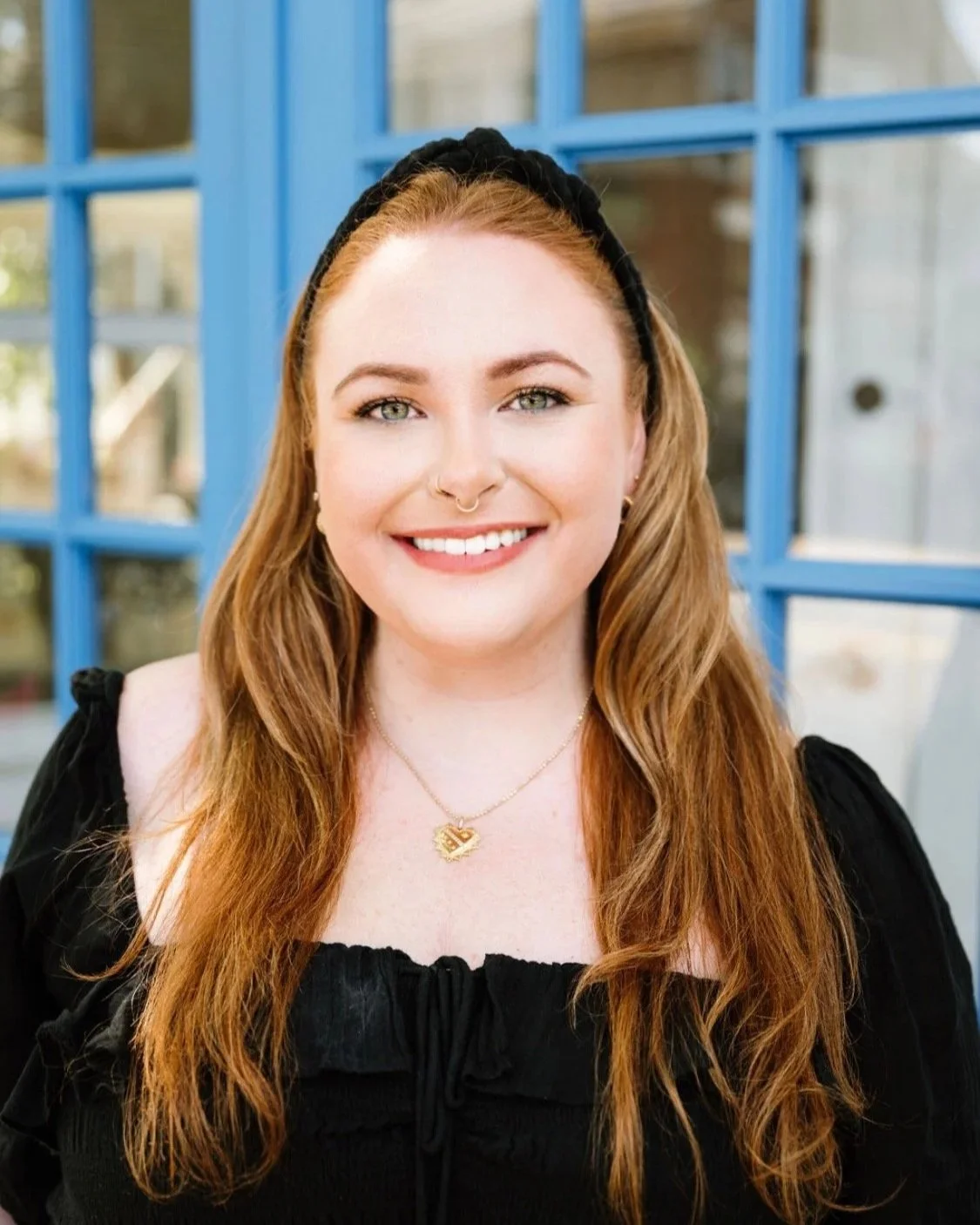 A young woman with long, wavy red hair, blue eyes, and a nose ring smiling outdoors in front of blue window frames.