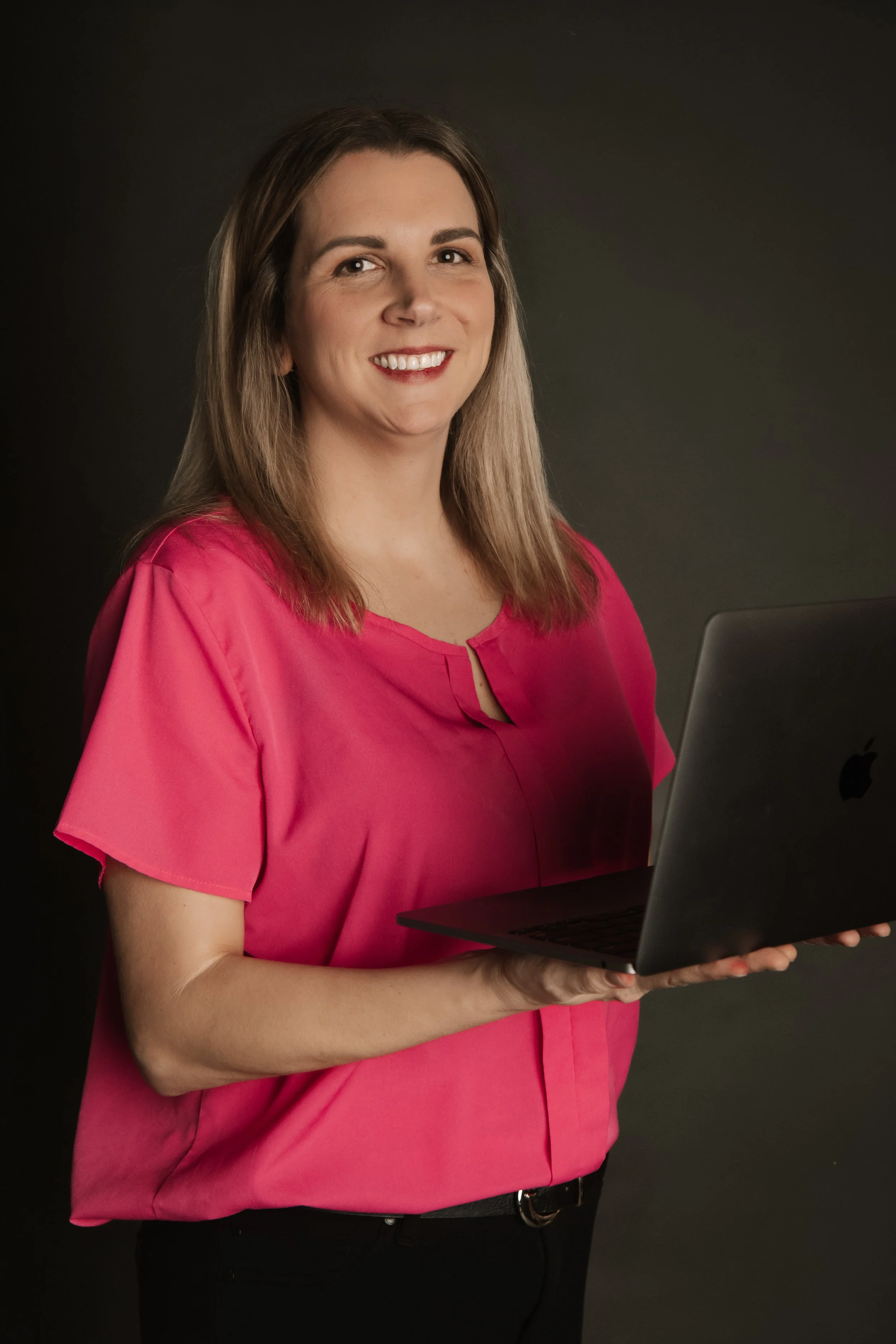 Woman with brown hair wearing a pink blouse holding a laptop and smiling