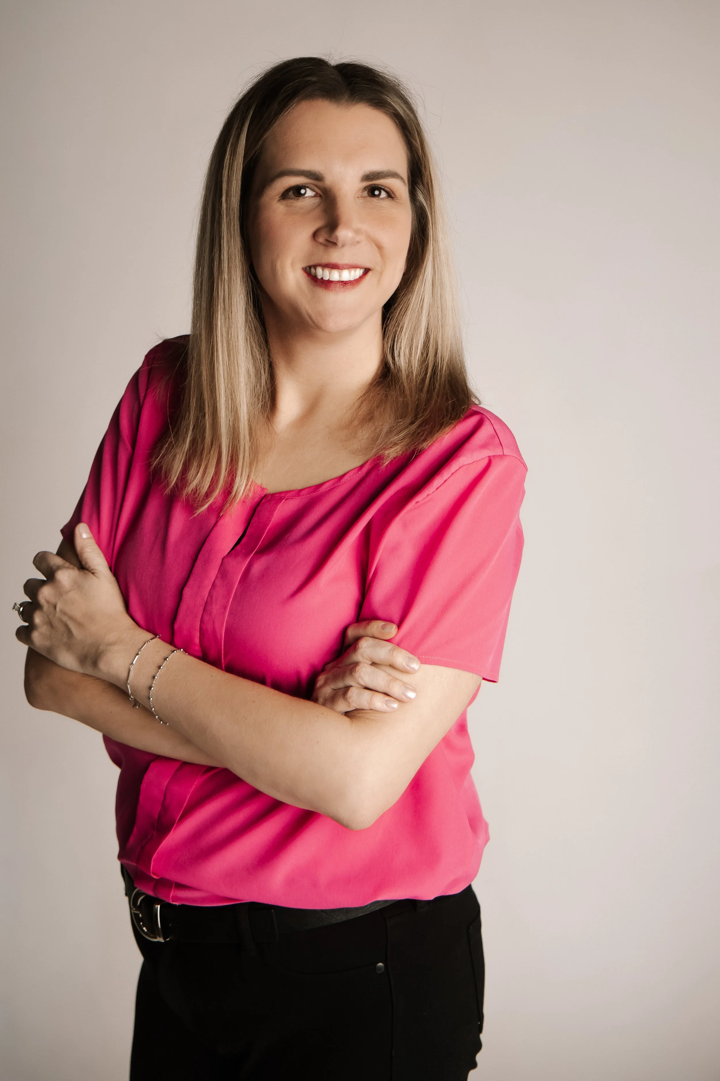 A woman with light brown hair wearing a pink blouse and black pants, standing with her arms crossed, smiling at the camera against a plain background.