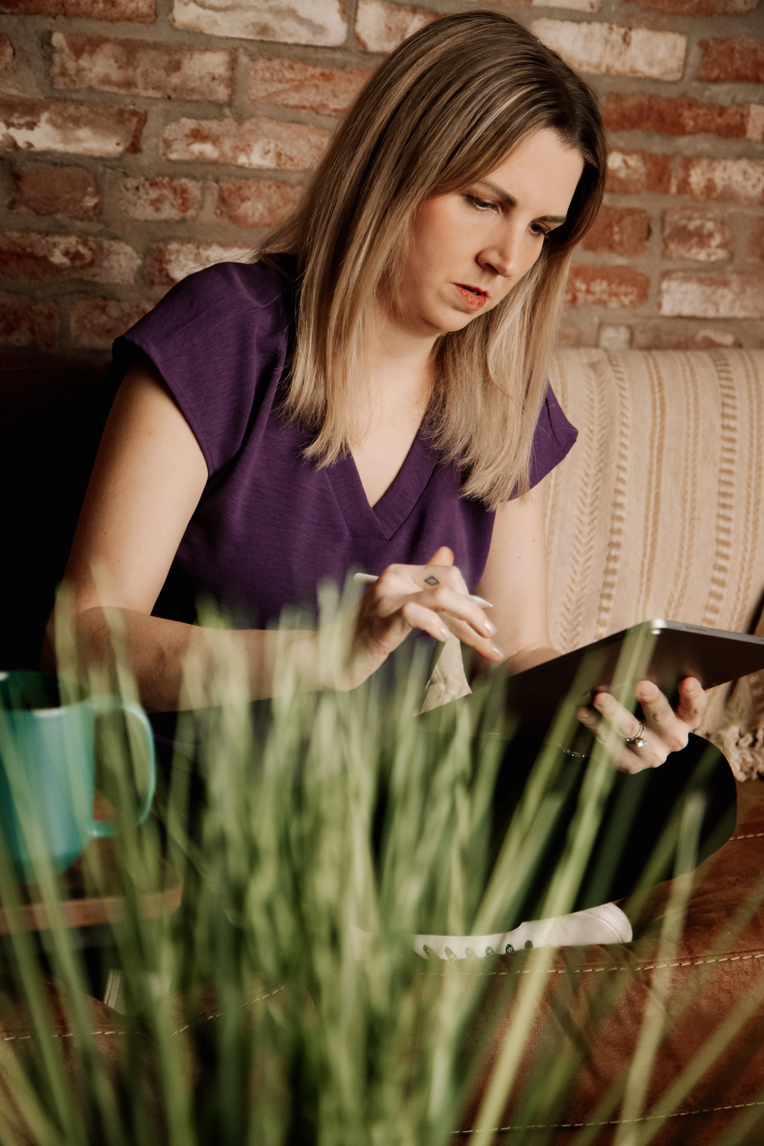 A woman with shoulder-length blonde hair, wearing a purple top, sits at a table with a brick wall in the background. She is focused on a tablet, holding a stylus in her right hand and touching the screen with her left hand. There is a green mug and some blurred greenery on the table.