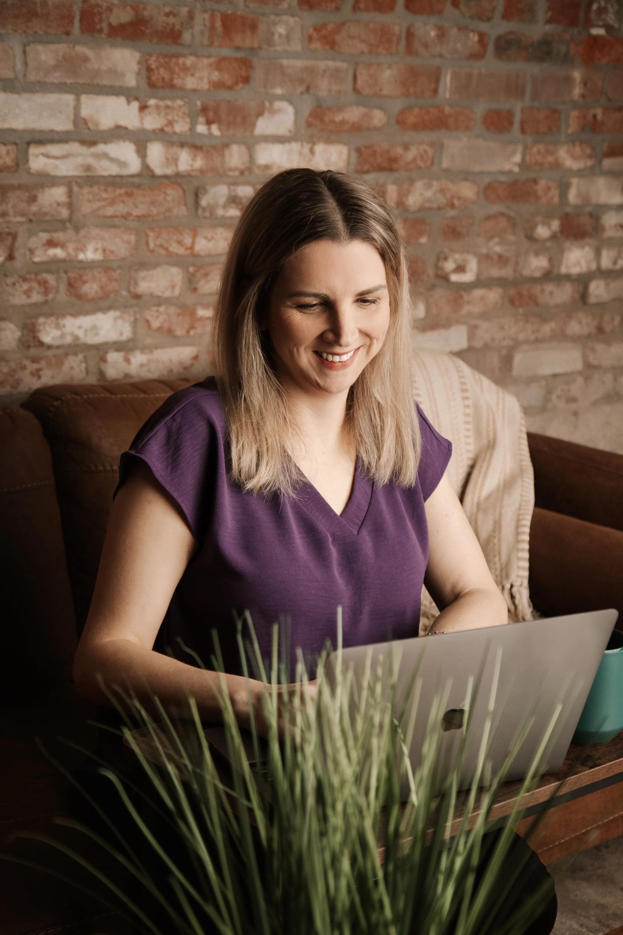 A woman with shoulder-length blonde hair wearing a purple shirt, sitting on a brown couch with a brick wall in the background, smiling while using a laptop on a wooden table. In the foreground, there's a plant and a teal cup.