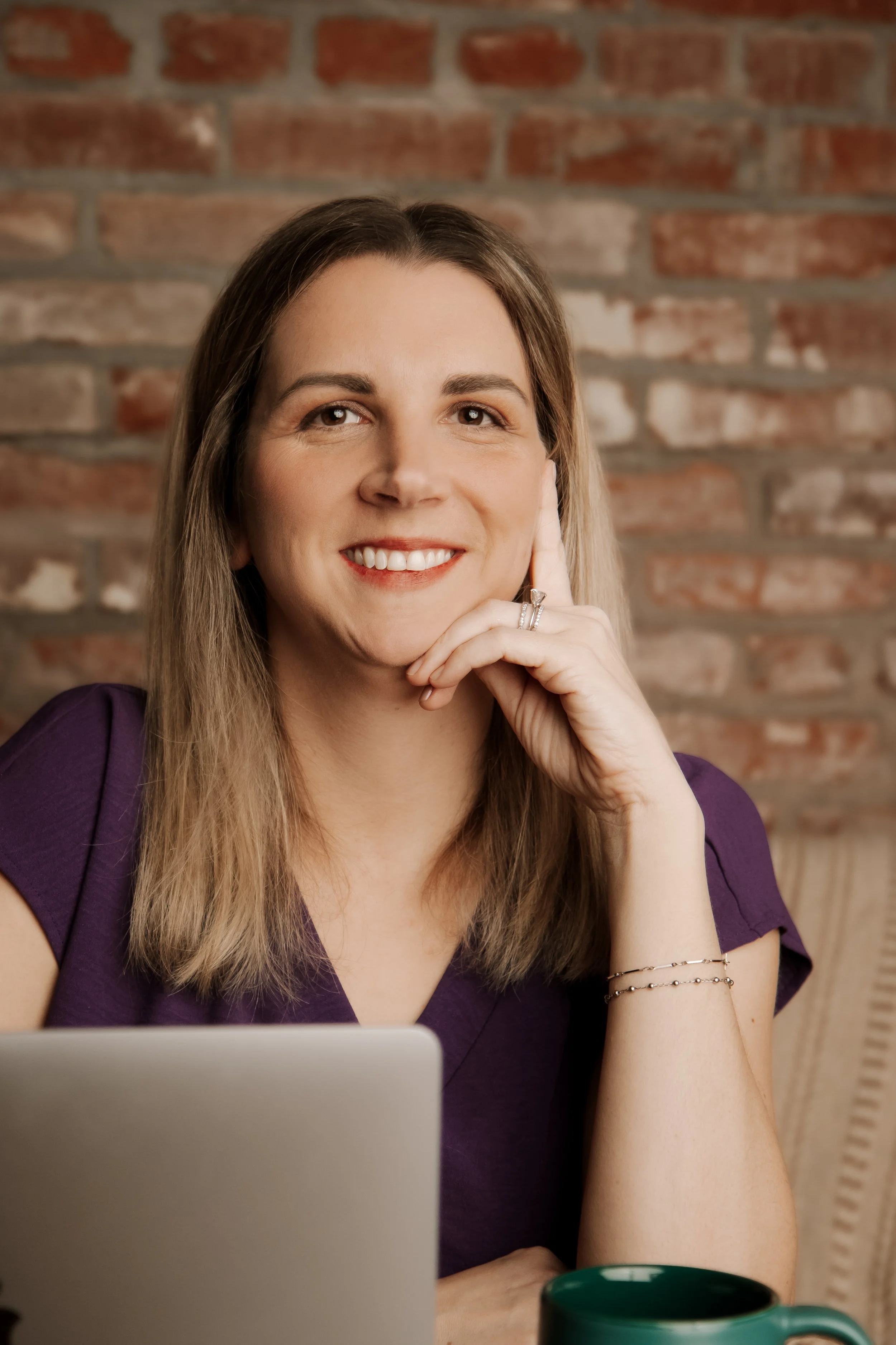 A woman with long blonde hair and a purple top, smiling with her hand resting on her face, sitting in front of a brick wall with a laptop and a green mug.