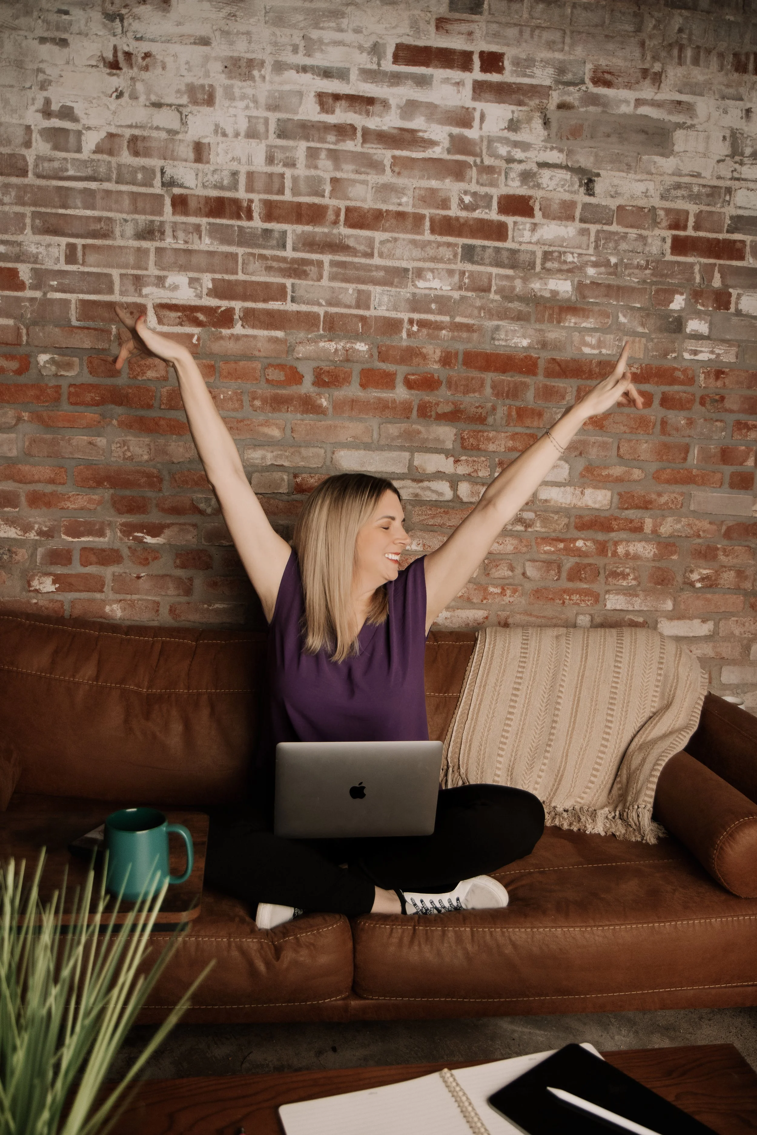 A woman sitting on a brown leather couch with a laptop, stretching with arms raised, smiling, in a cozy room with a brick wall background.