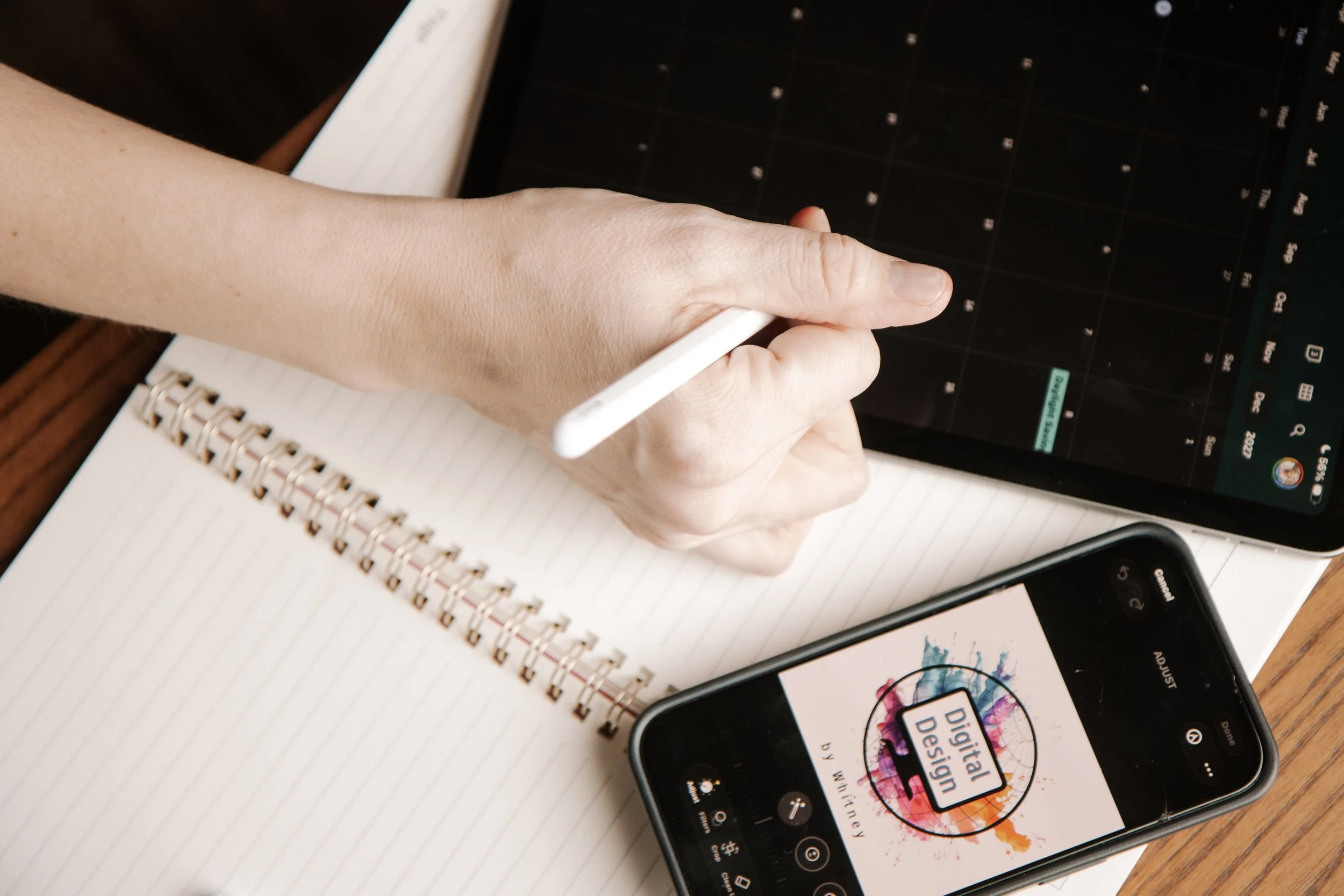 A person holding a stylus and writing in a spiral notebook while using a tablet with a digital calendar or planner. A smartphone with a colorful logo reading 'Digital Design' is also on the desk.