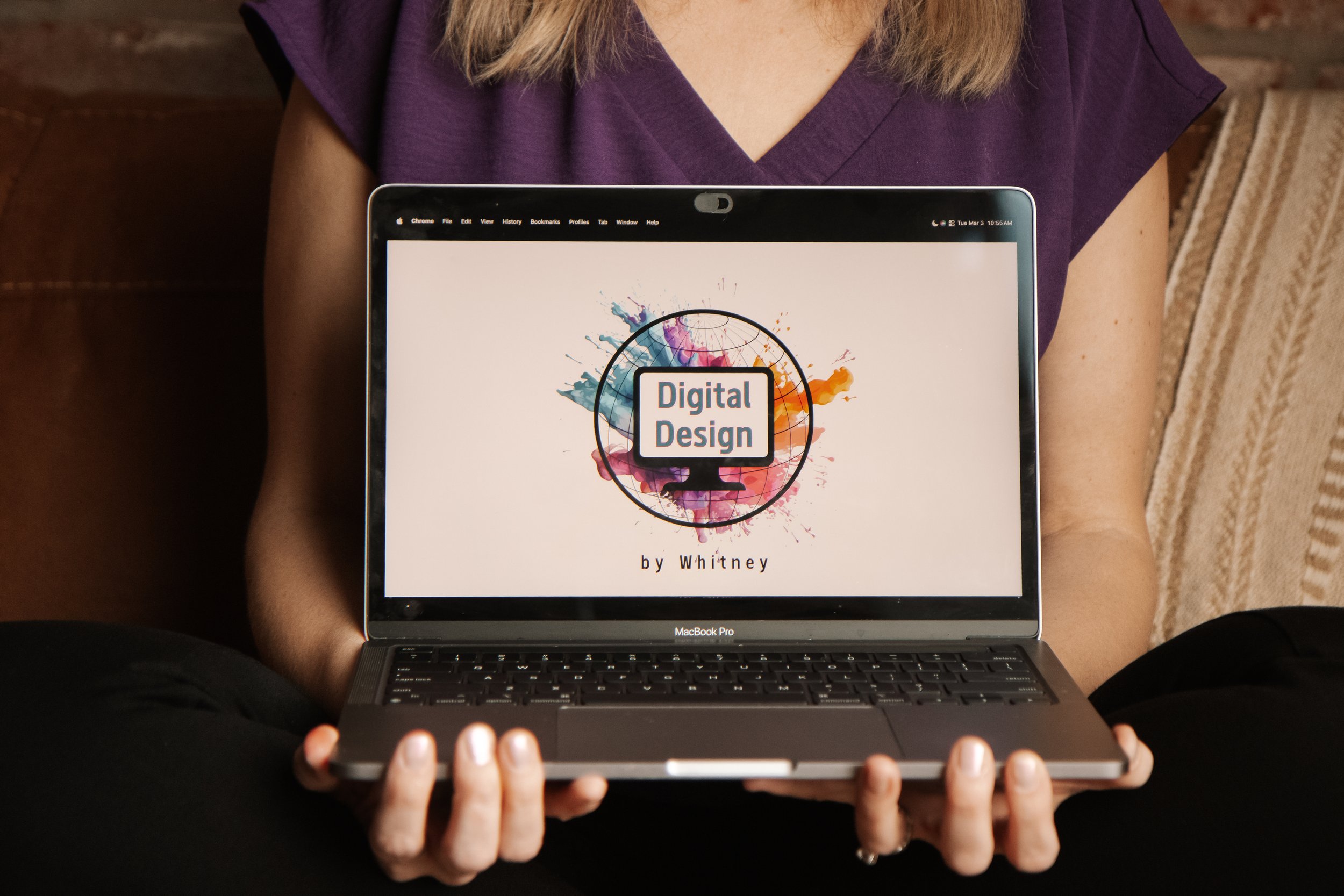 Person sitting cross-legged holding a MacBook Pro displaying a digital design project with a colorful splash background and the text 'Digital Design by Whitney.'