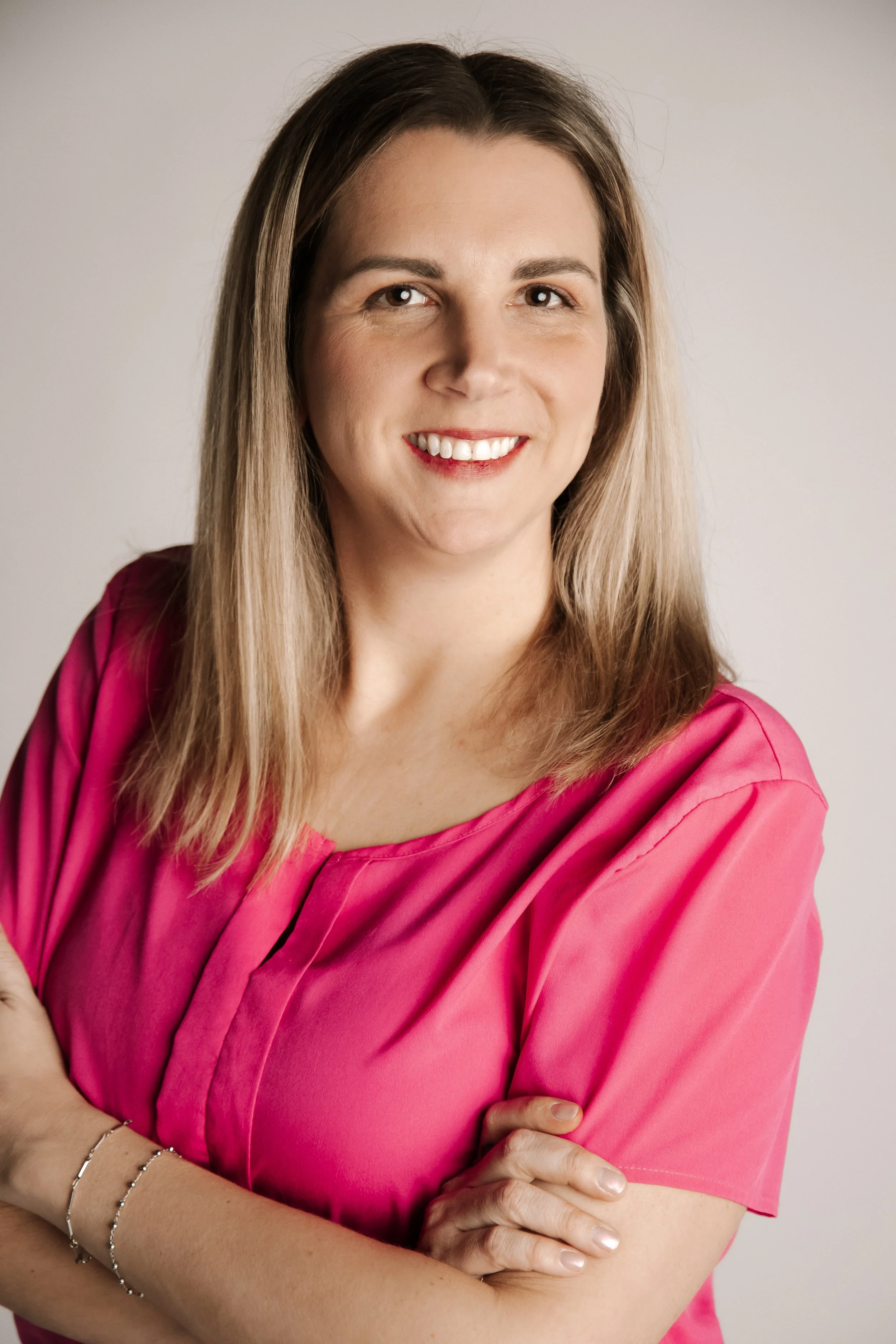 A woman with shoulder-length blonde hair, smiling, wearing a pink top, with her arms crossed.