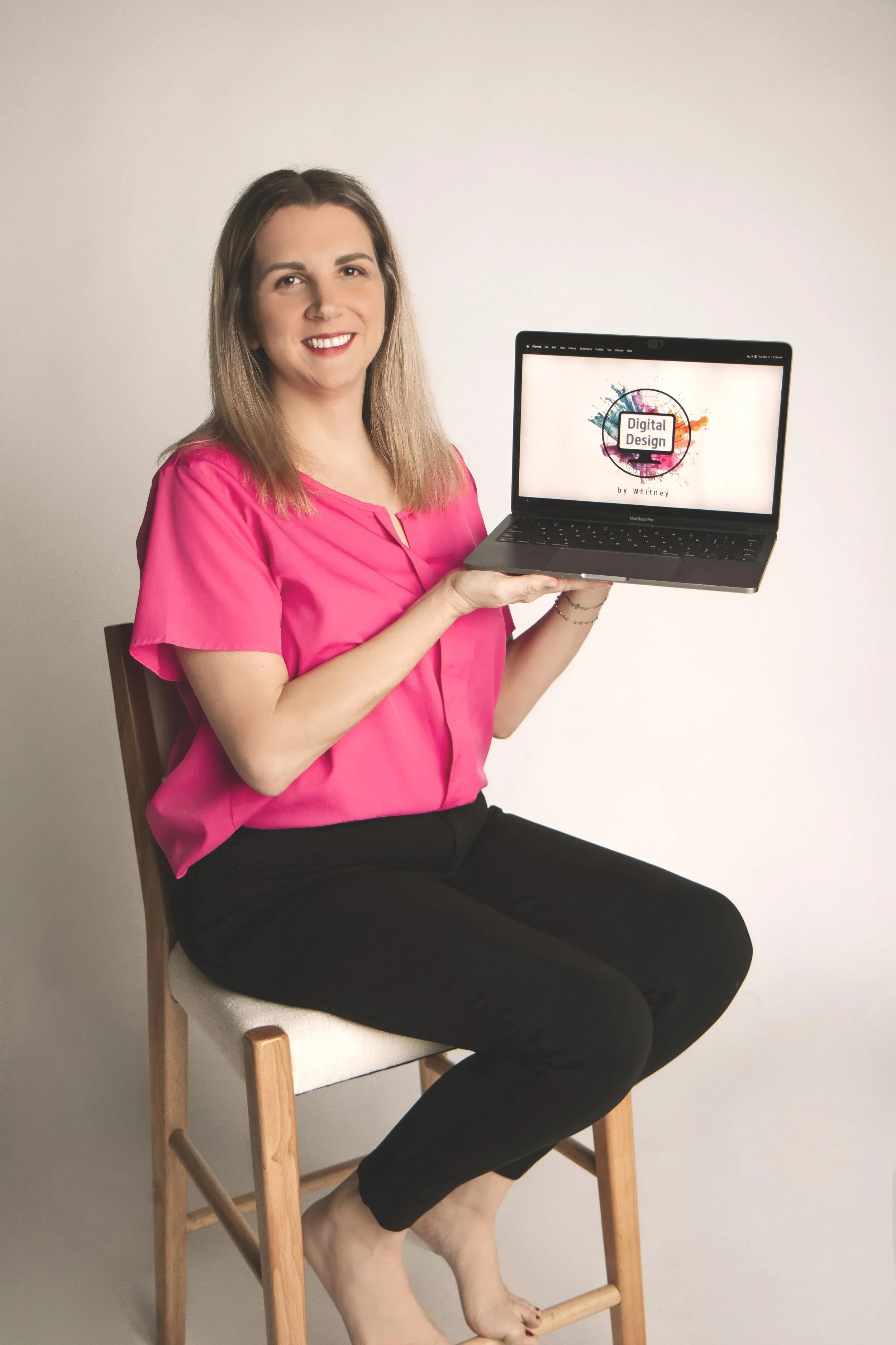 A woman with shoulder-length brown hair, wearing a pink blouse and black pants, sitting on a wooden chair with no shoes, holding a laptop displaying a colorful 'Digital Design' graphic.