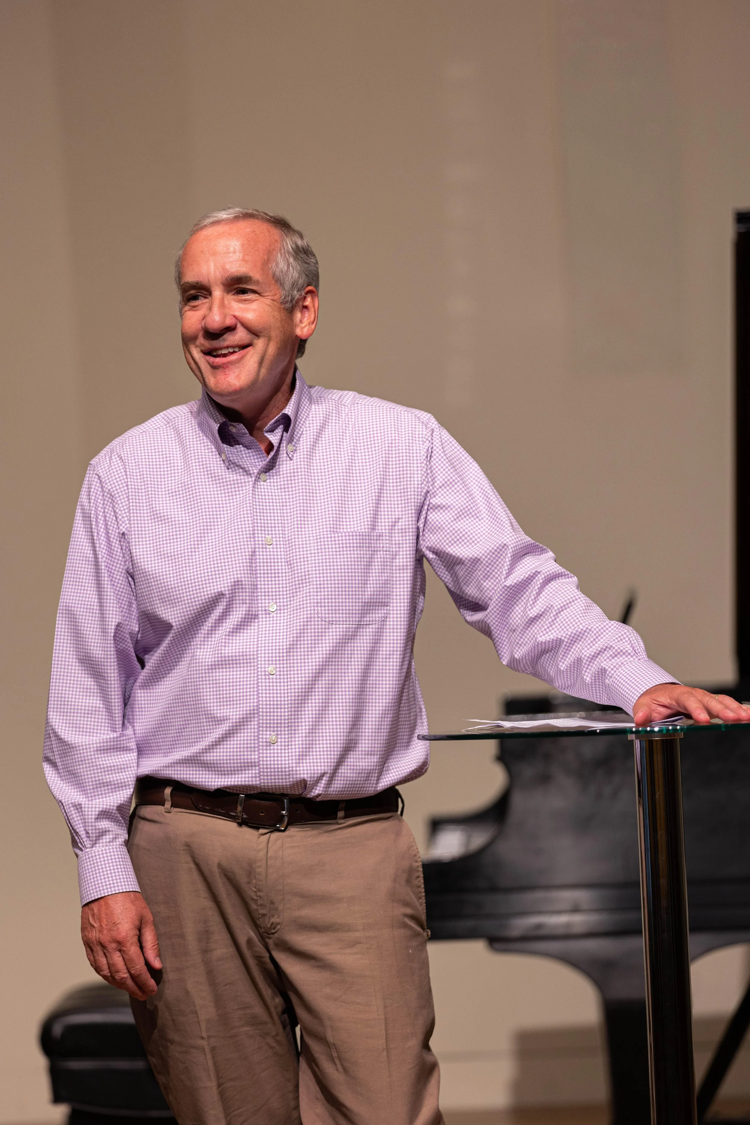 A middle-aged man with gray hair smiling, wearing a light purple checkered shirt and khaki pants, standing next to a piano.