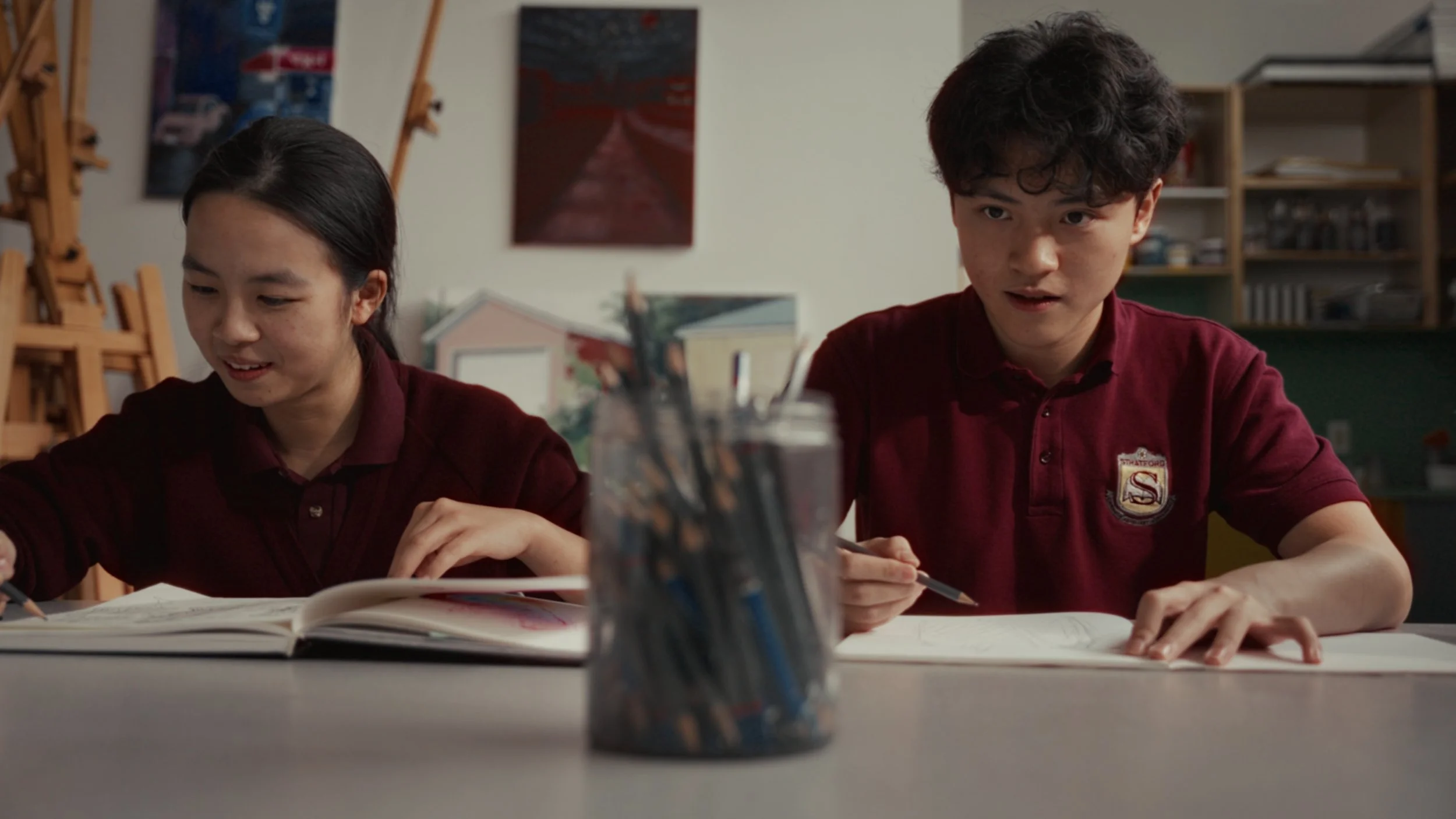 Two students sitting at a table, working in their notebooks, with a jar of colored pencils in the foreground, in a classroom setting.