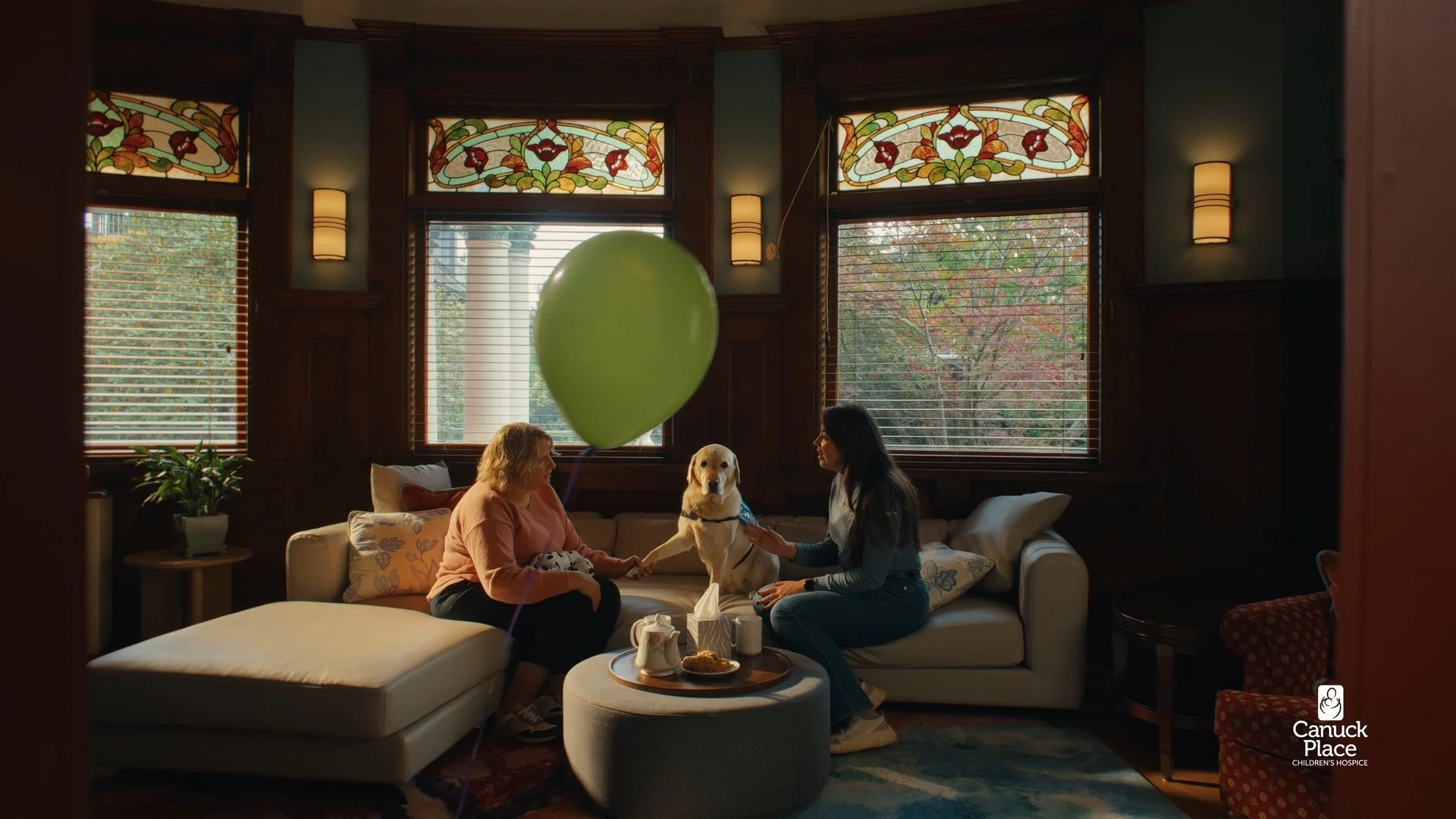 Two women and a service dog sitting on a beige sofa in a cozy living room with stained glass windows, wooden paneling, and a small table with tea and snacks.