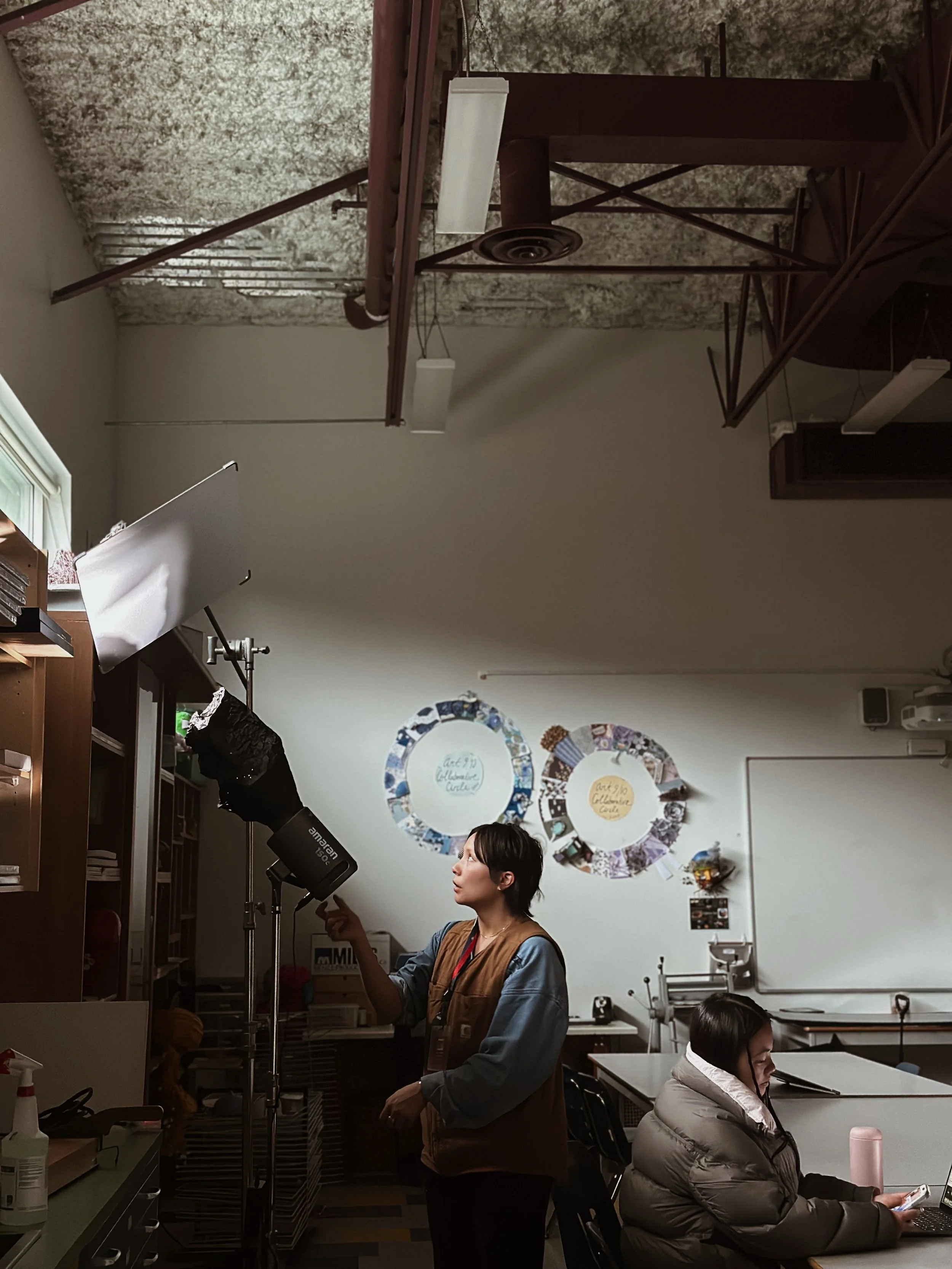 An indoor classroom scene with two women. One woman is adjusting studio lights, and the other is sitting at a desk using a laptop. Decorations on the wall include two large wreaths with writing.