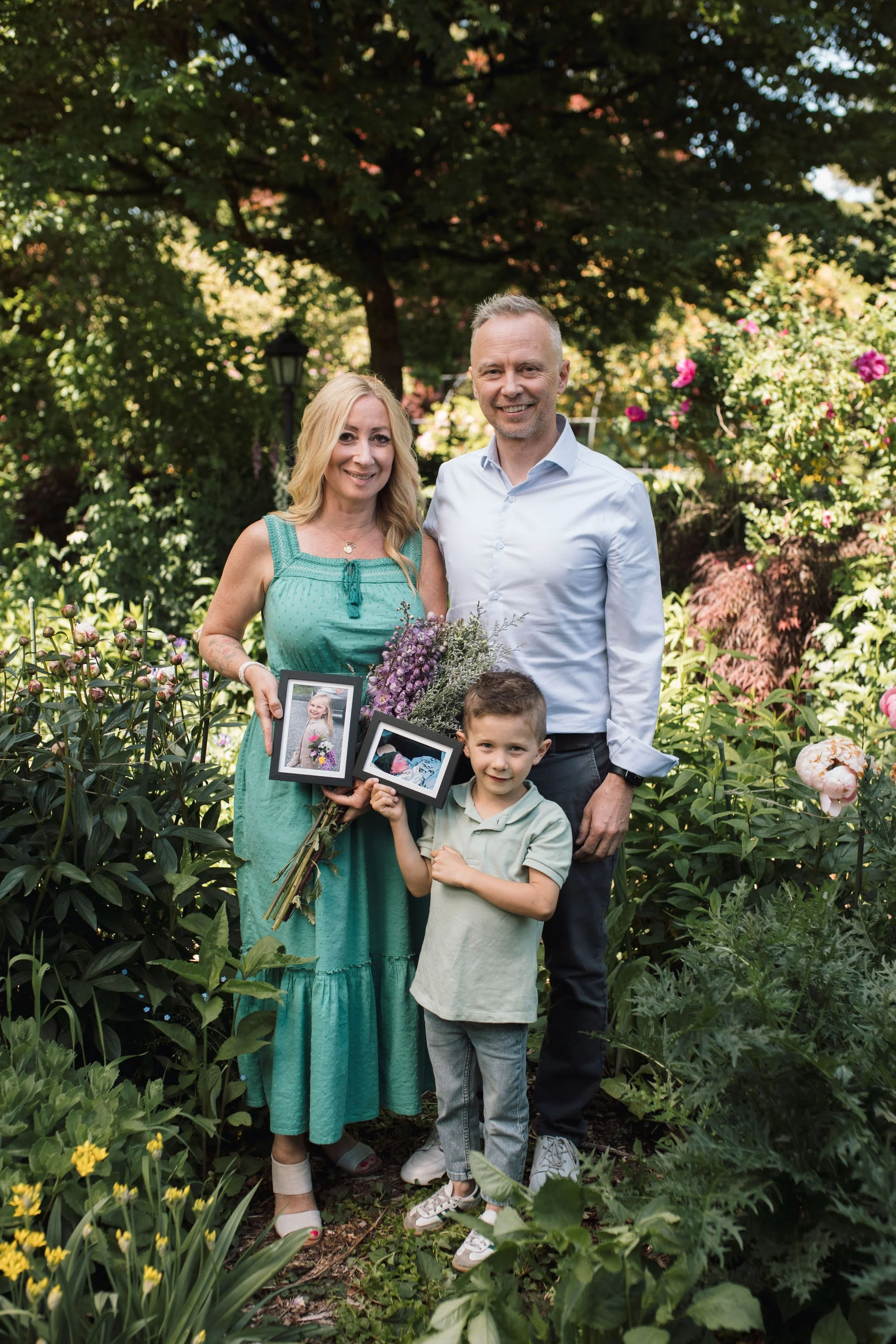 A family of three, a woman, a man, and a young boy, standing in a lush garden with flowers and trees, smiling for a photo. The woman holds framed photographs and a bouquet of flowers.