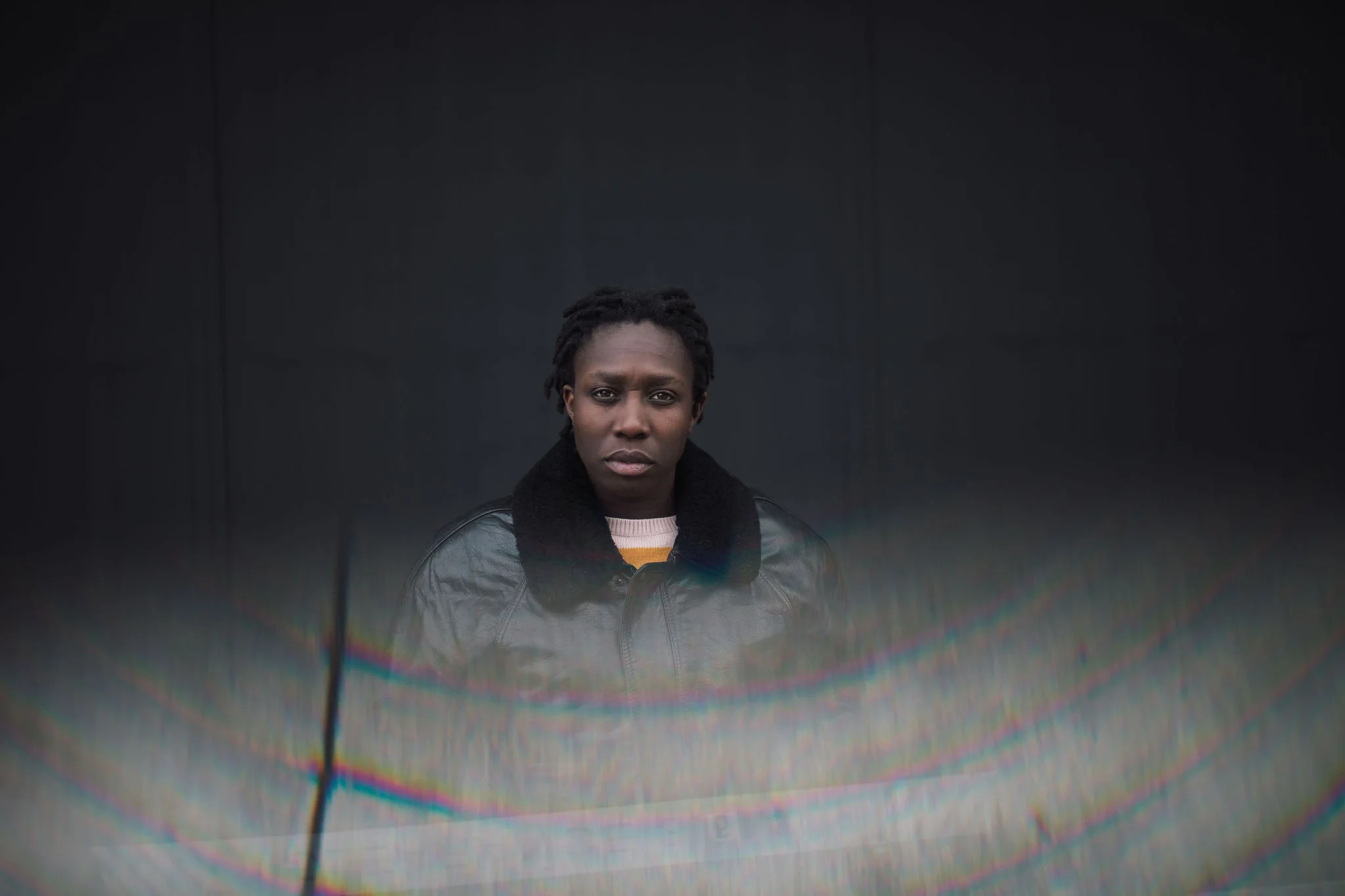 A woman with short dreadlocks wearing a leather jacket with a fur collar, standing in front of a black background, with a rainbow-colored lens flare across the lower part of the image.